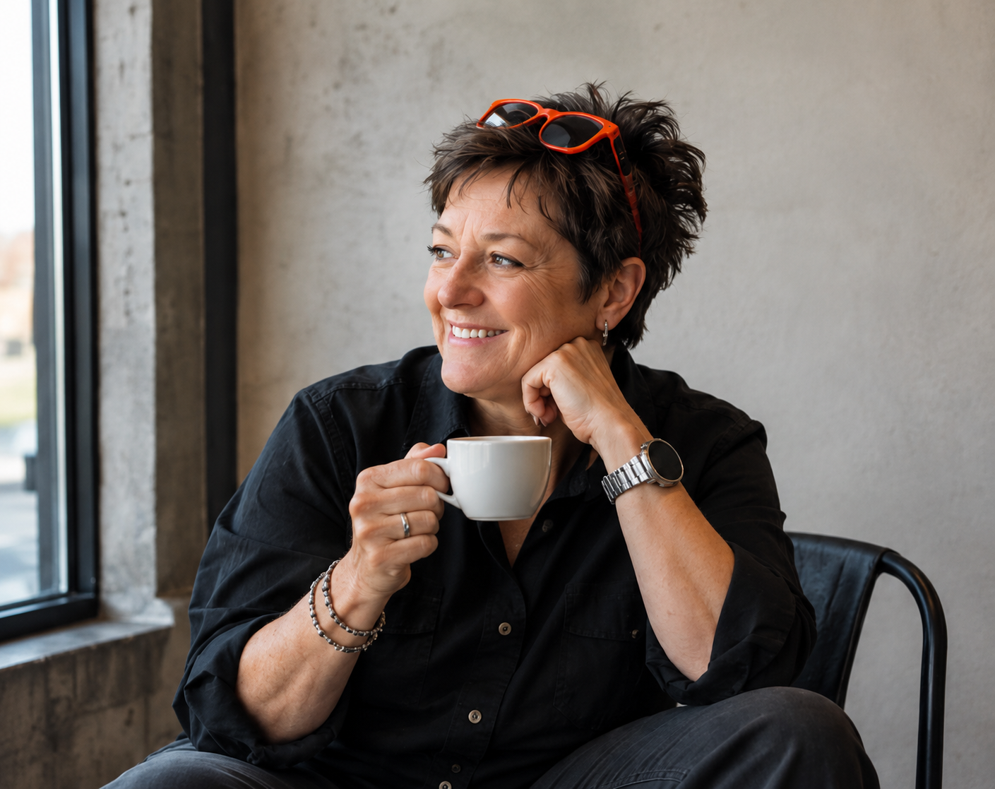 A middle-aged woman with short brown hair sitting near a window, smiling and holding a white coffee cup, wearing black clothing, glasses on her head, and jewelry, in a modern indoor setting.