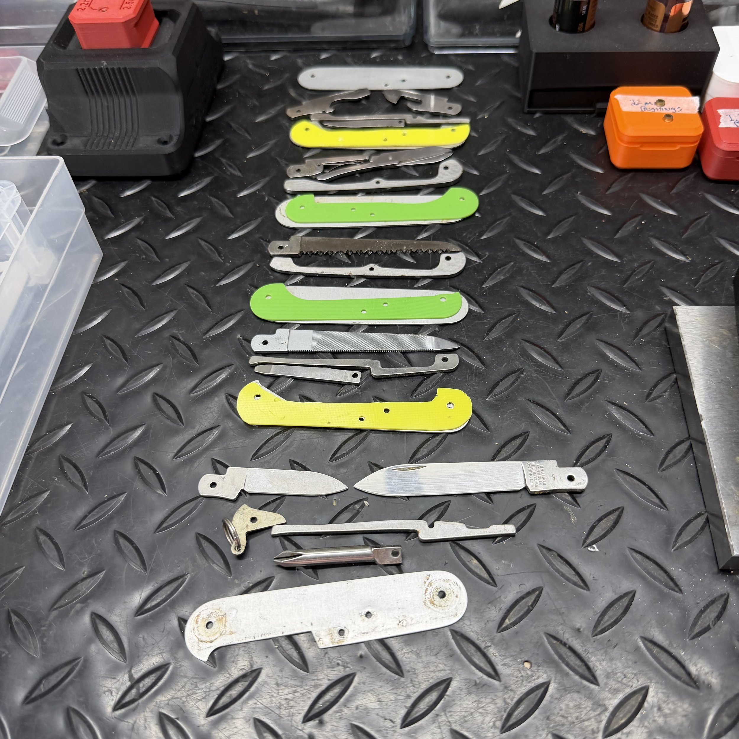 Various metal razor blades arranged in a row on a black textured surface, with some blades having bright yellow and green painted ends. In the background, there are plastic containers and small boxes, some with labels. The surface has a diamond plate pattern.