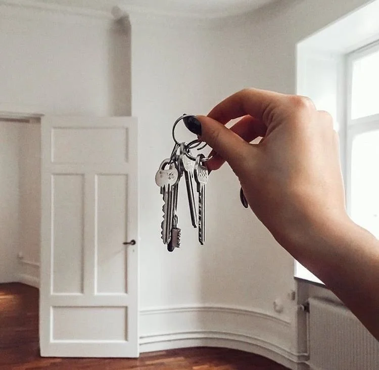 Person holding up a set of keys inside a bright room with white walls, open door, wood floor, and large window.