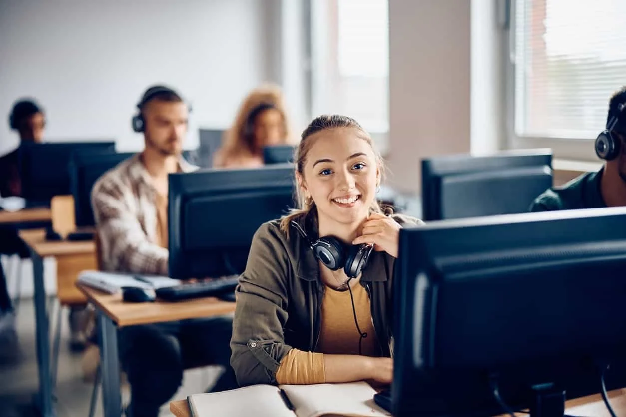 A young woman smiling at the camera in a classroom or computer lab, surrounded by peers working on desktop computers, all wearing headphones.