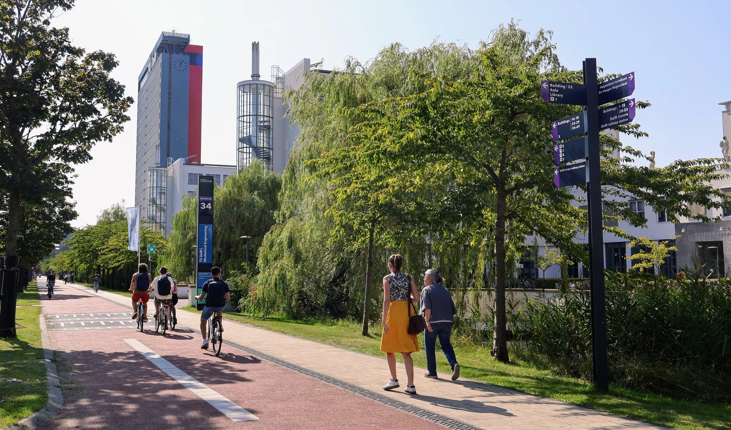 People riding bicycles and walking on a sidewalk in an urban park scene with trees and modern buildings in the background.