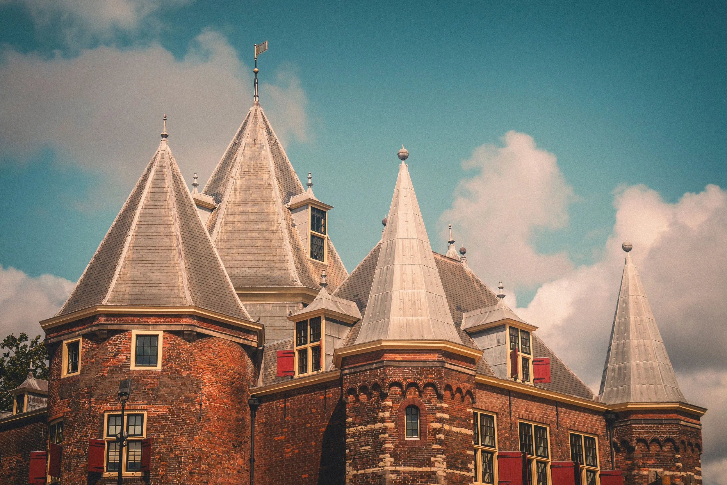 A historic red brick building with pointed, slate-gray rooftops and conical towers, set against a partly cloudy sky.
