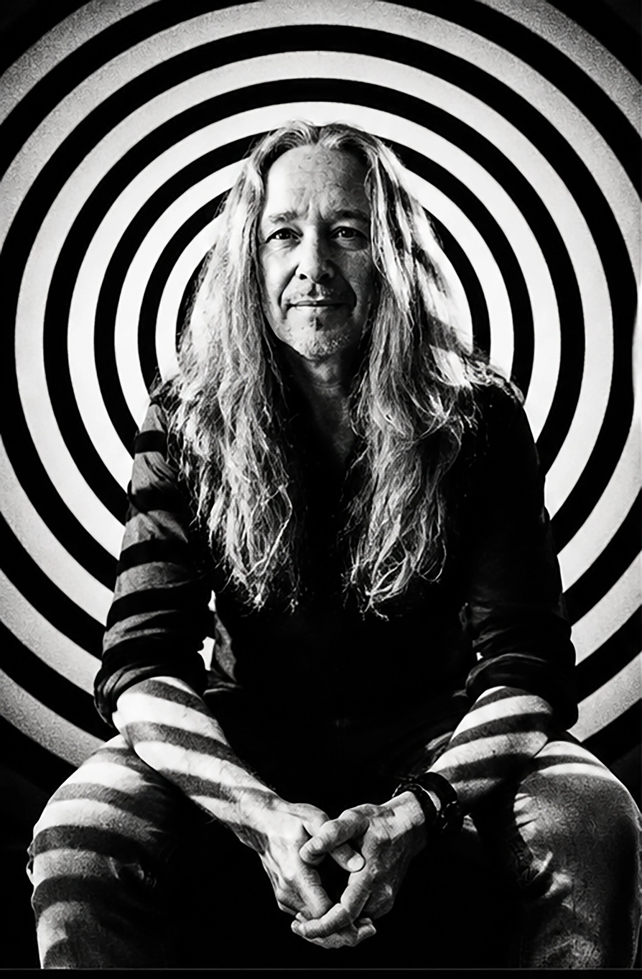 Black and white portrait of a man with long hair, sitting with hands clasped together, in front of a black and white concentric circle background. Op-Art, hypnotic psychedelic mesmerizing vibe and effect.
