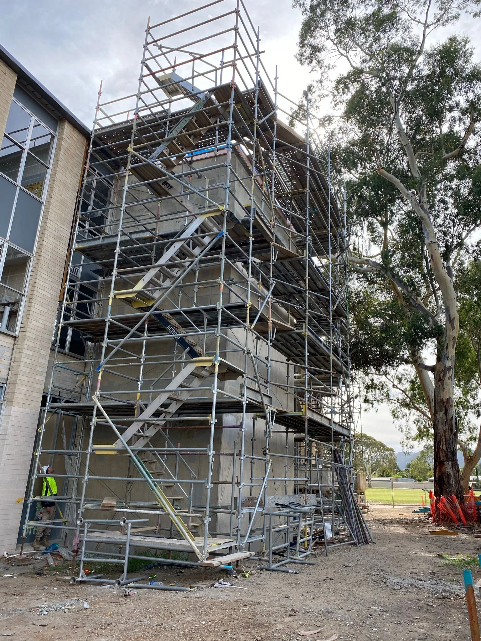 Construction site with scaffolding around a multi-story building, workers in safety vests, and a large tree nearby, under a cloudy sky.