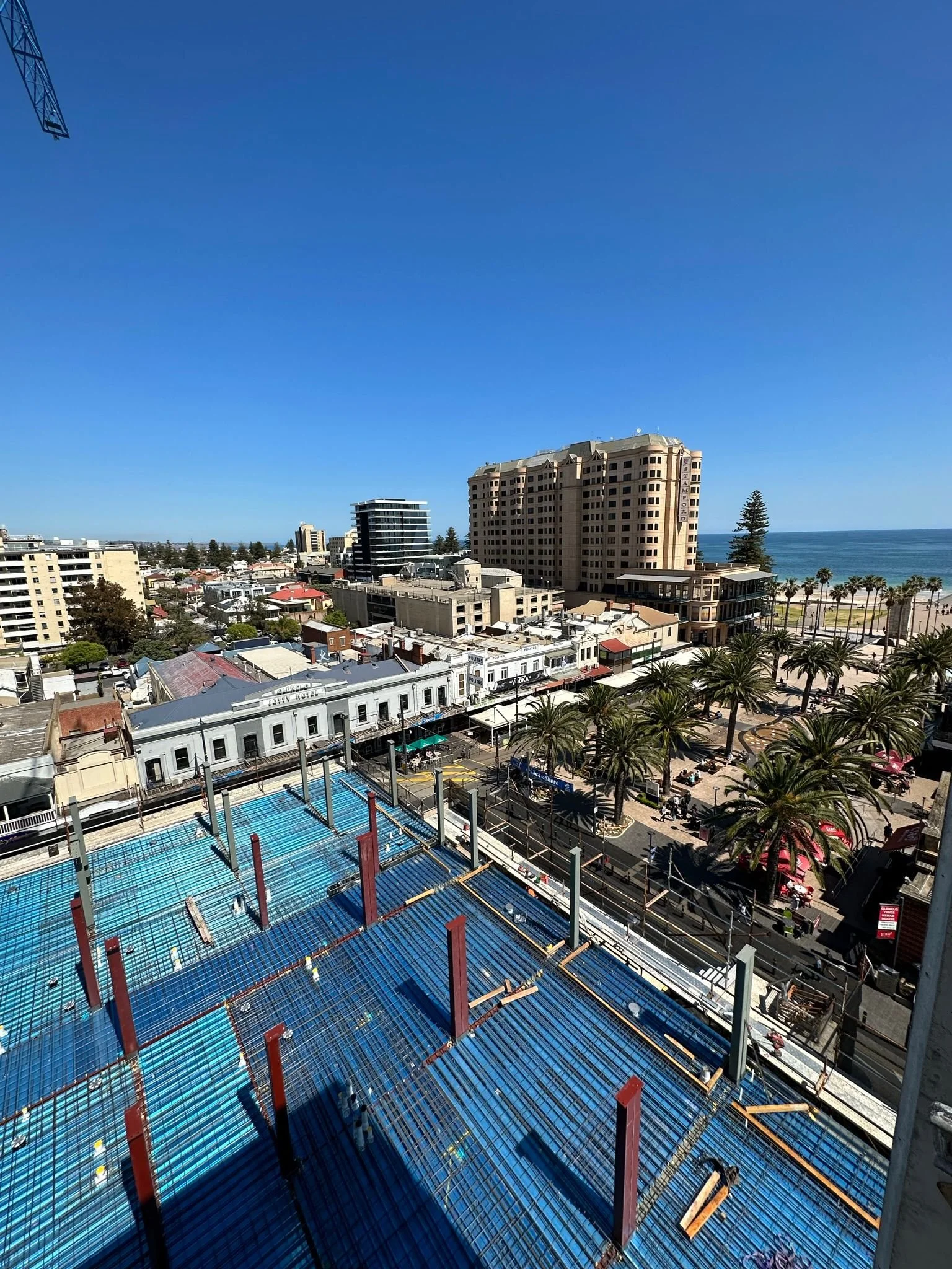 A cityscape view with a construction site in the foreground, featuring blue and red steel framework, surrounded by buildings, with the ocean and a clear blue sky in the background.