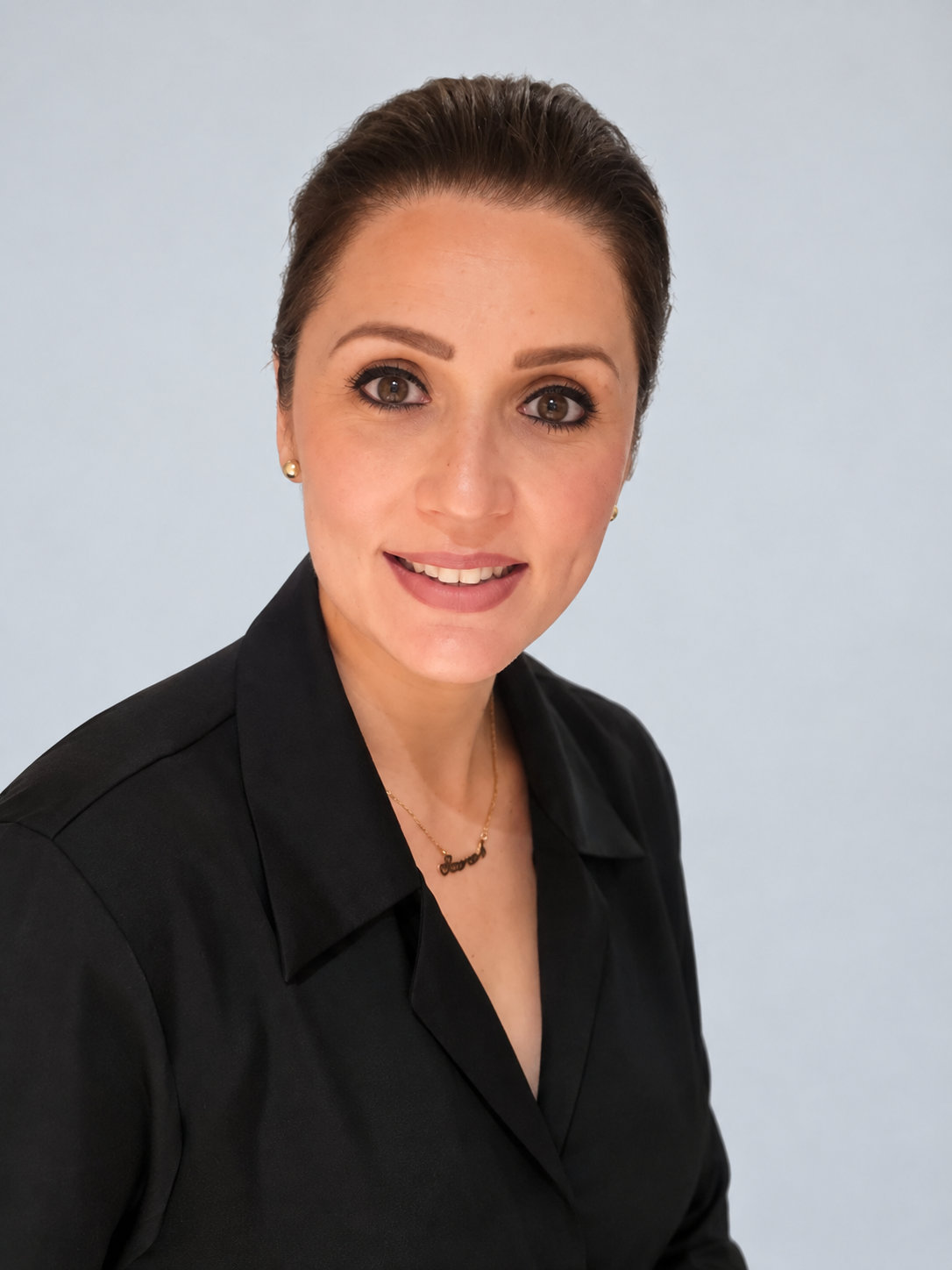 A woman with short brown hair, wearing a black shirt, pearl earrings, and a gold necklace with a name pendant, smiling at the camera against a light blue background.