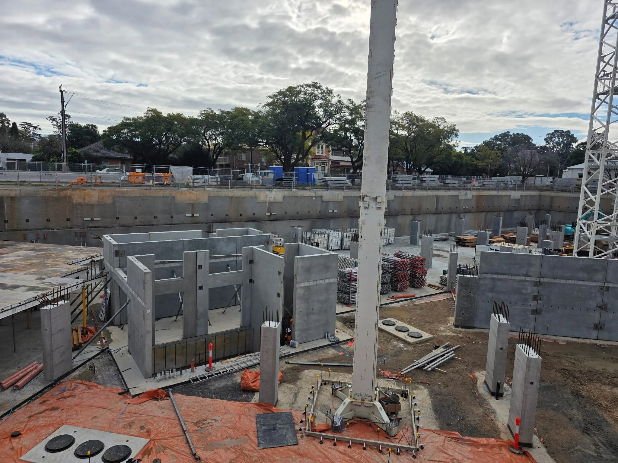 Construction site with building framework, concrete walls, metal scaffolding, and construction materials, with cloudy sky and some trees in the background.