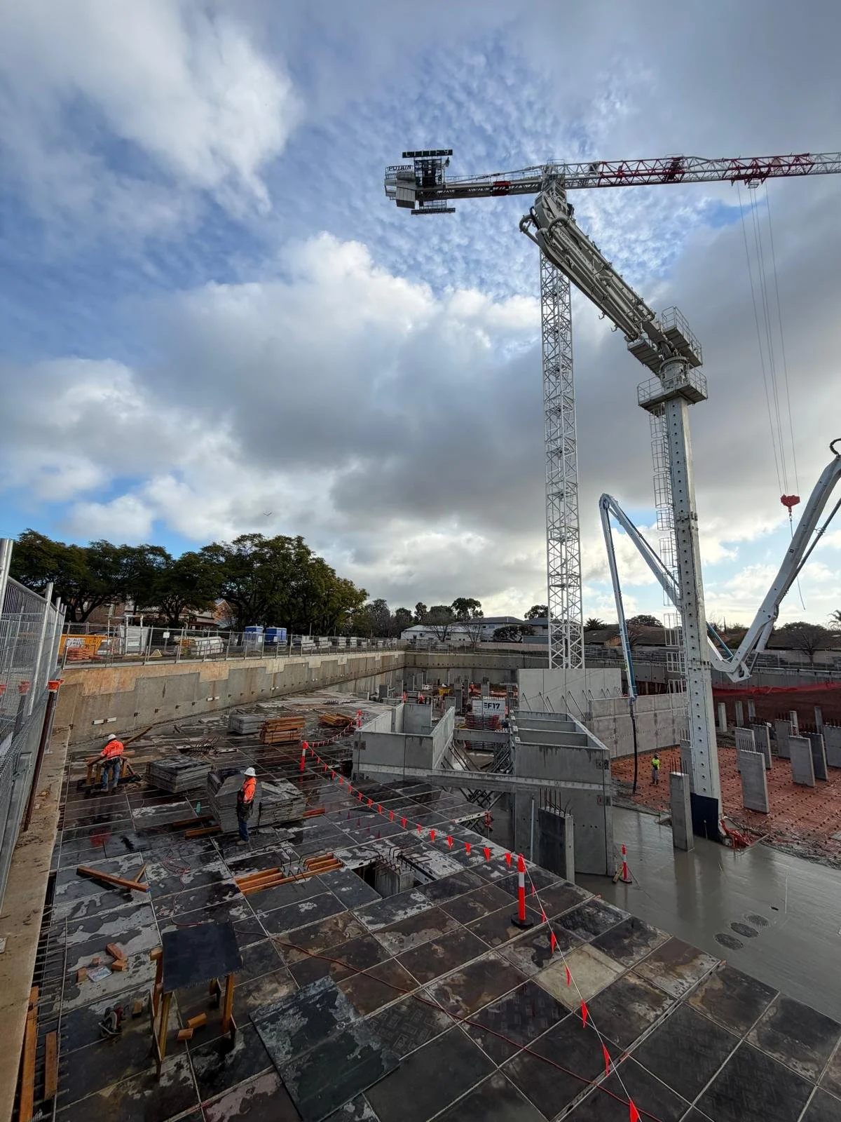 Construction site with a large tower crane, workers wearing safety vests and helmets, and concrete foundation being laid with scaffolding.