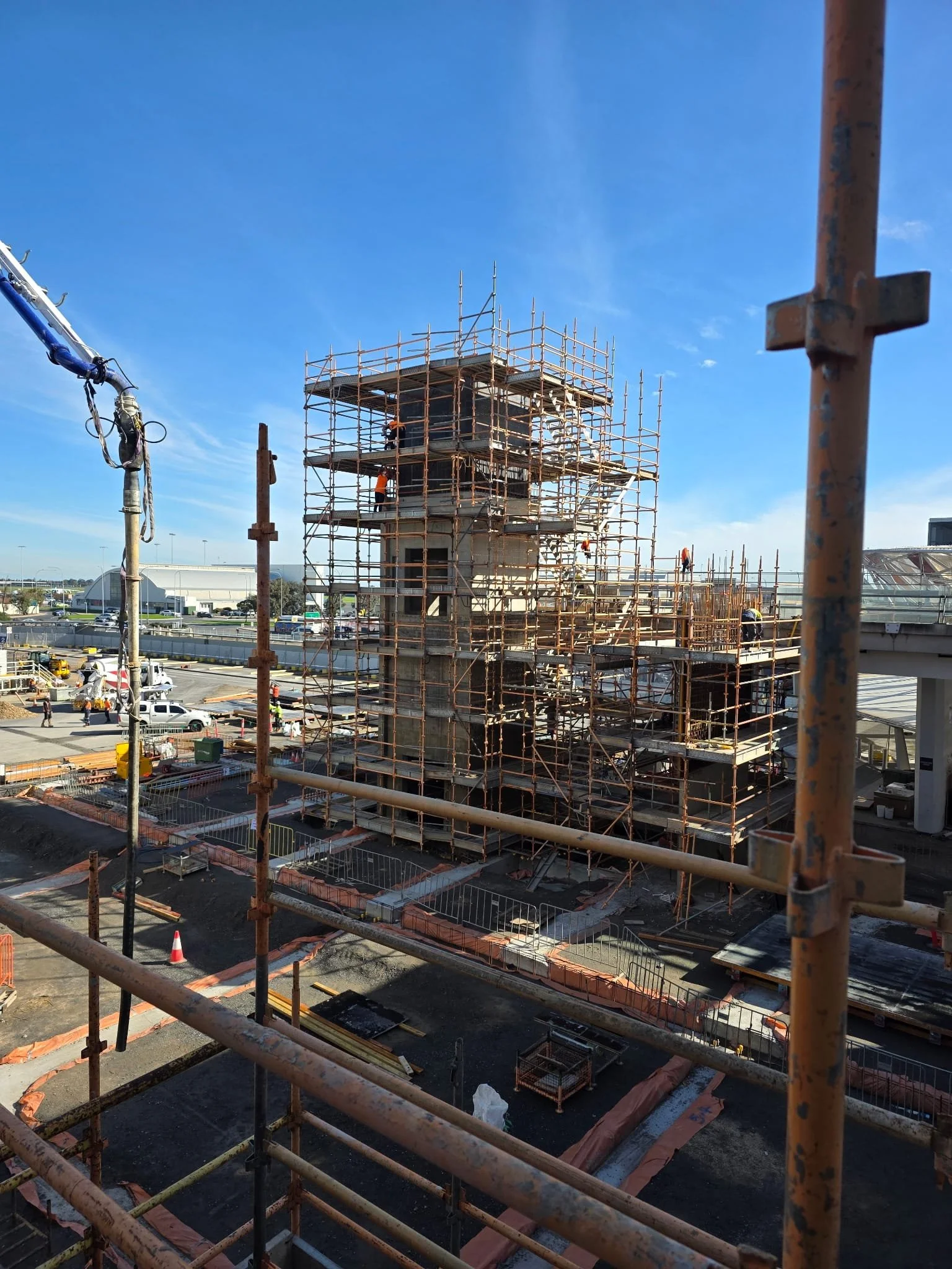 Construction site with scaffolding around a multi-level building structure, partially built, with workers and construction equipment, under a blue sky.