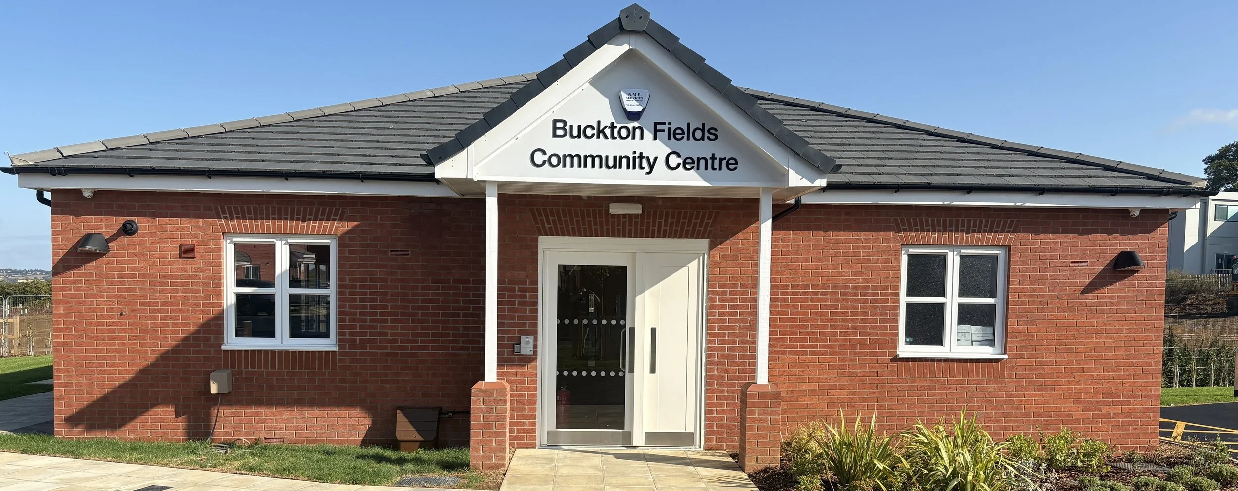 Front view of Buckton Fields Community Centre, a single-story brick building with a gray tiled roof, white signage, and two windows, set against a clear blue sky.