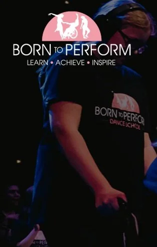 A person wearing a black shirt with a logo that says BORN TO PERFORM DANCE SCHOOL, standing in front of a dark background with a sign that has the same logo and slogan.