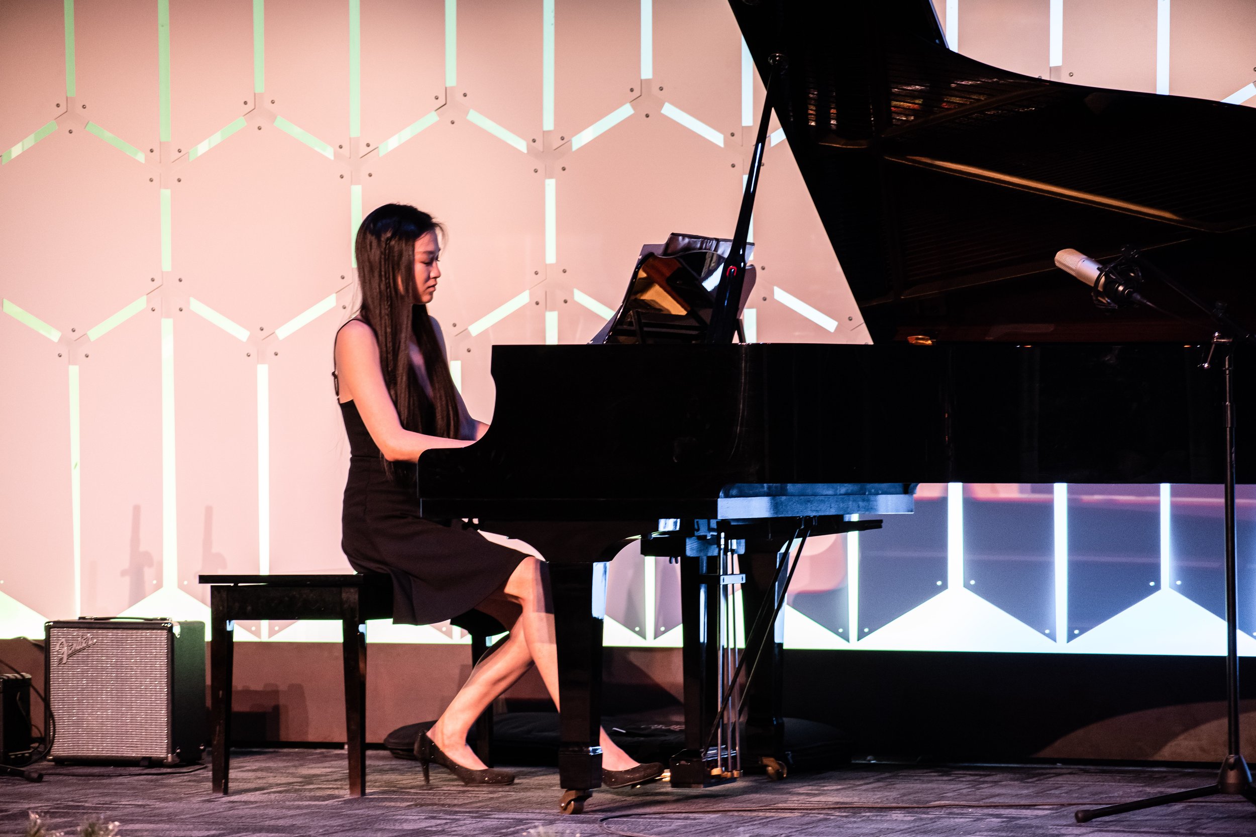 A woman with long dark hair wearing a black dress, playing a grand piano on a stage with a modern geometric light wall in the background.