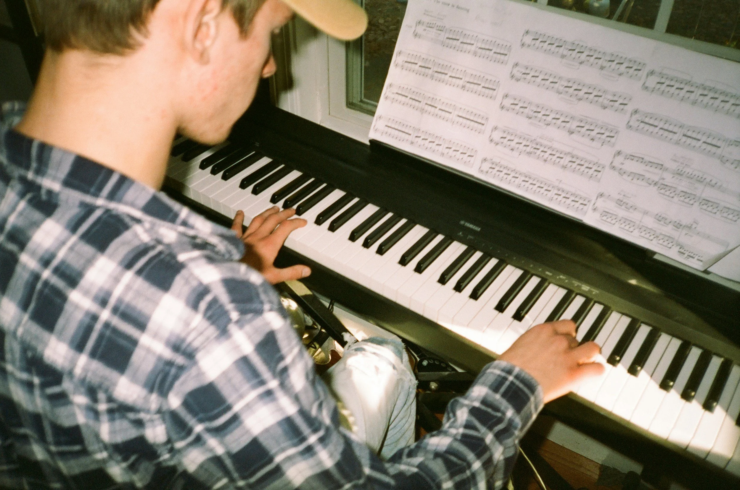 A person wearing a plaid shirt is playing a black upright piano, reading sheet music placed on the music stand, in a room with a window in the background.
