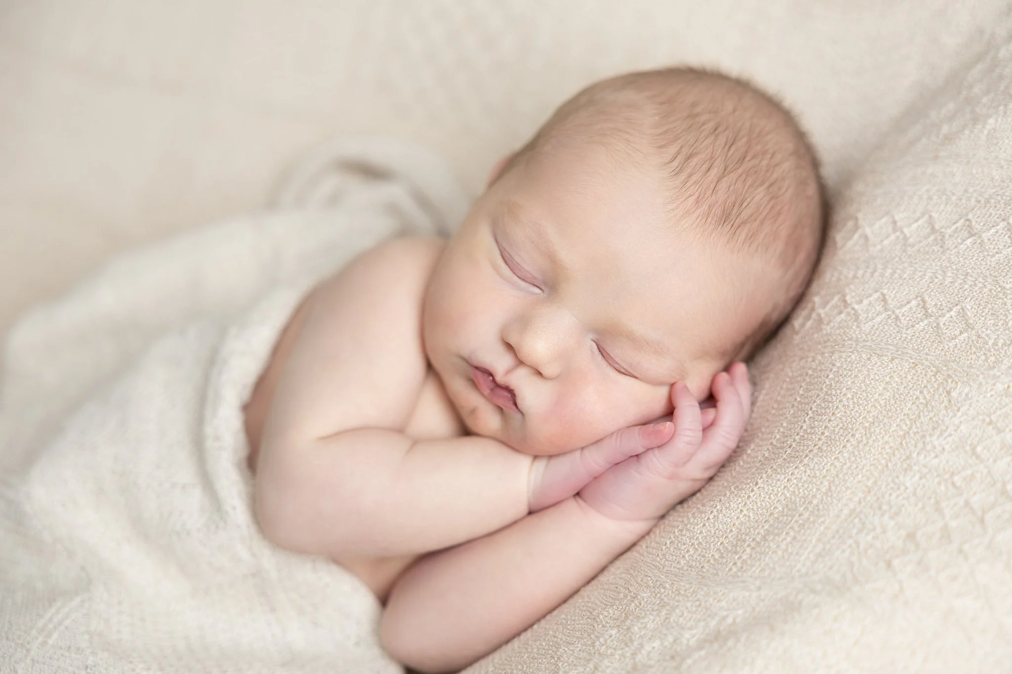 Close-up of a sleeping newborn baby with a peaceful expression, lying on a soft, beige textured blanket.