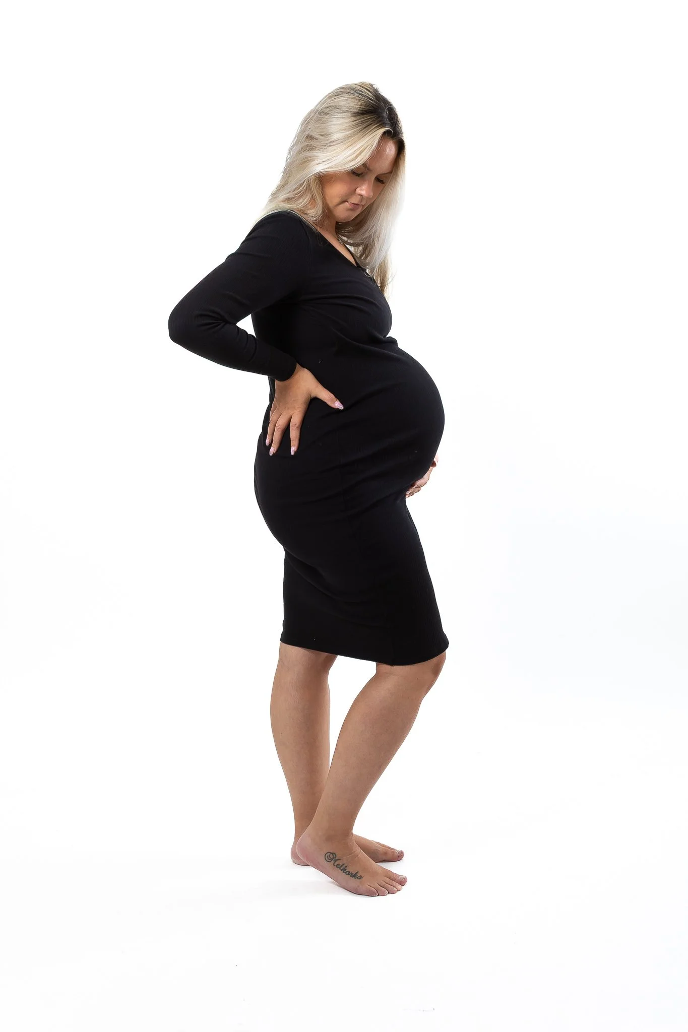 Pregnant woman in black dress standing against a white background with her hands on her back and her head slightly bowed, showing her pregnancy.