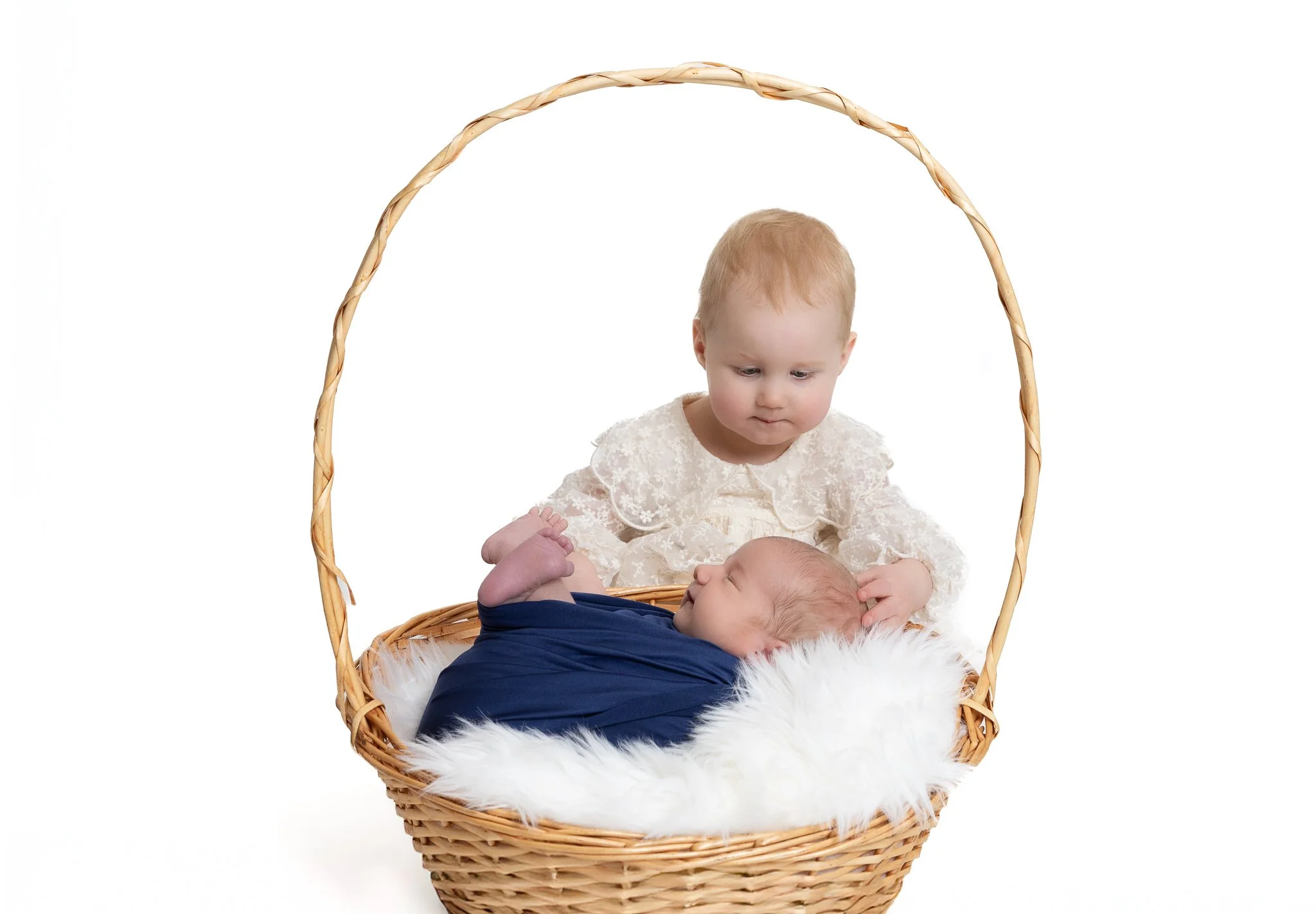 A young child and a baby in a wicker baby carriage with a white fur lining against a white background.