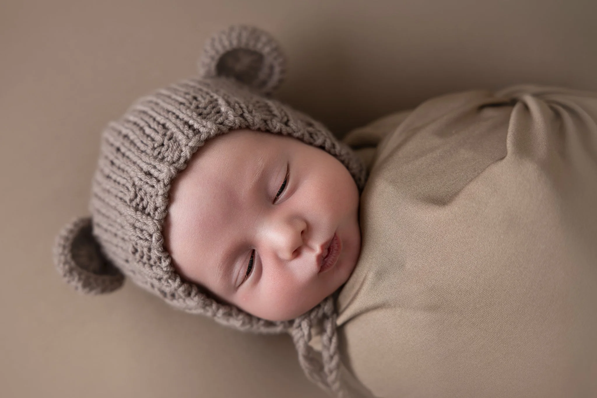 Close-up of a sleeping baby wearing a knitted hat with bear ears and a beige outfit, lying on a neutral background.