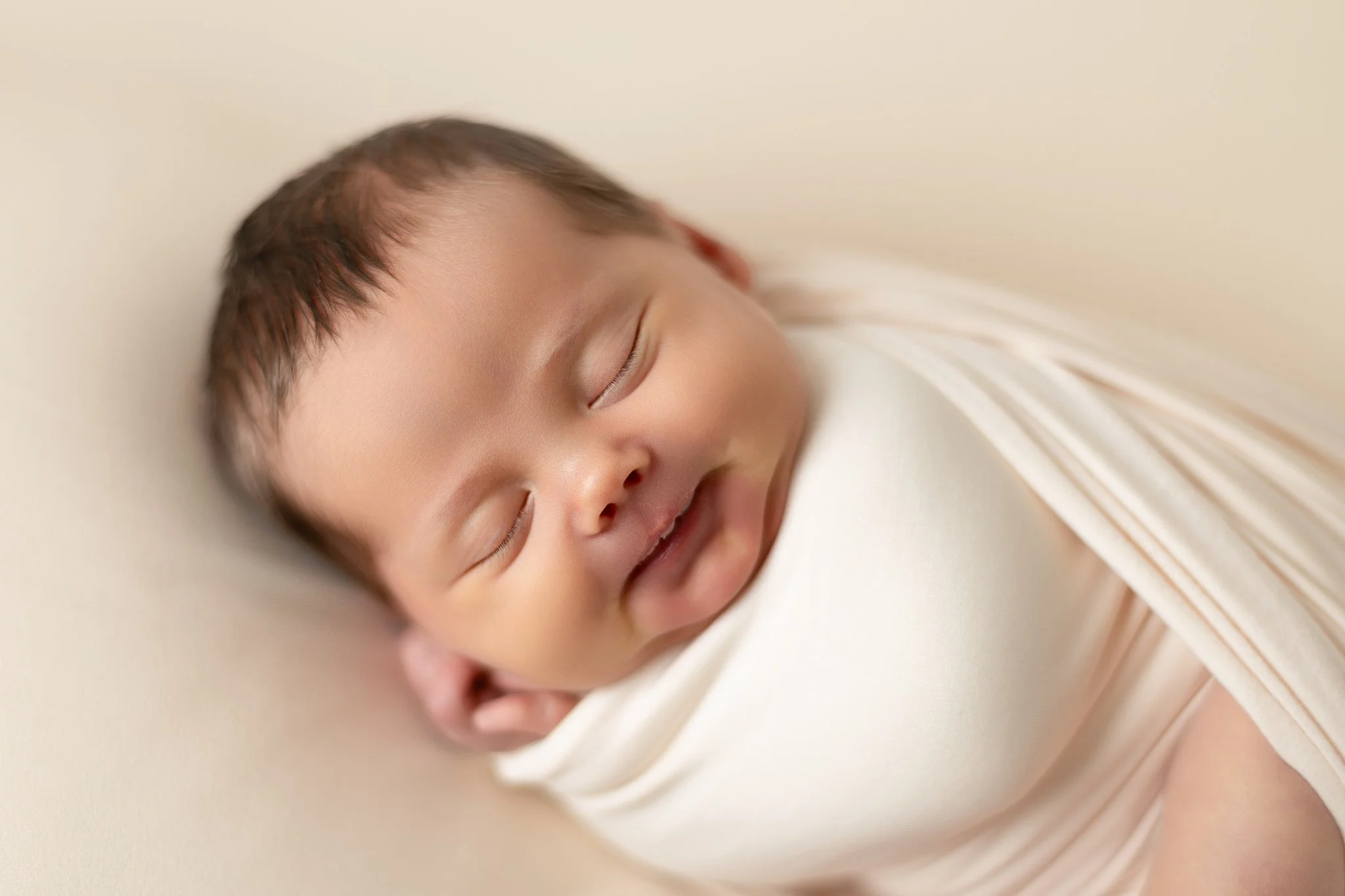 Close-up of a sleeping baby with closed eyes, lying on a soft surface wrapped in a light cream blanket.