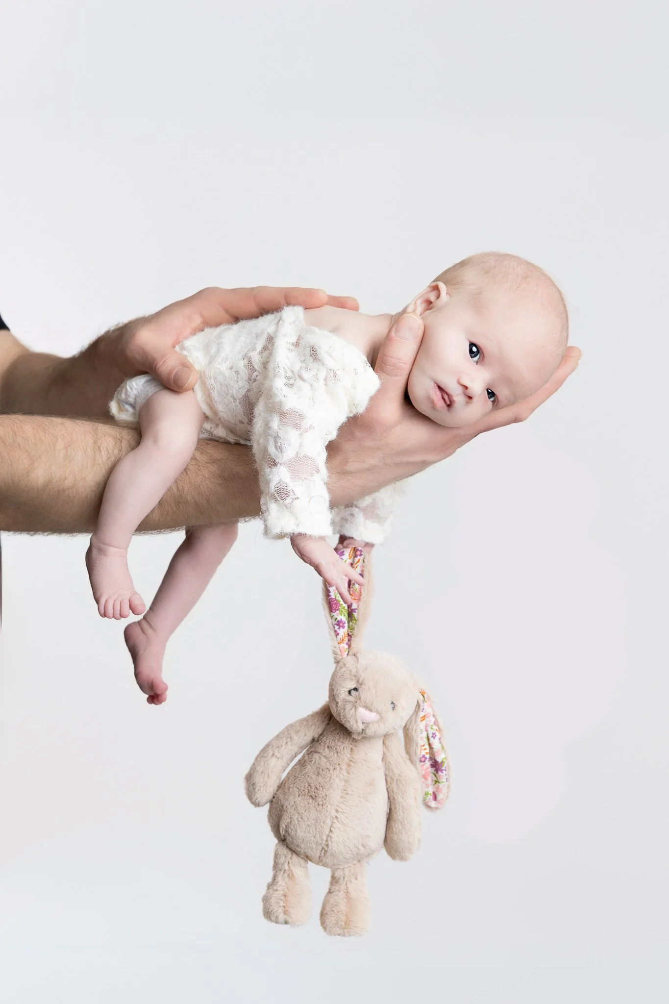 A baby's head resting on an adult's hand, with the baby lying on its side while holding a stuffed animal. The baby has a curious expression, large eyes, and light-colored hair. The stuffed animal is a beige plush bunny hanging down, with a floral rib