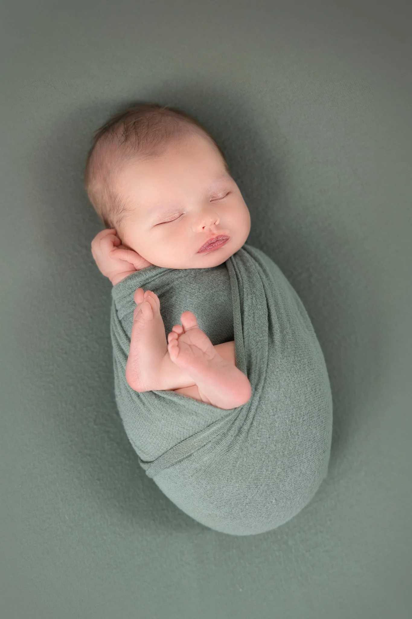 A sleeping baby wrapped in an olive green blanket, lying on a matching green surface, with the baby’s face and hand visible.