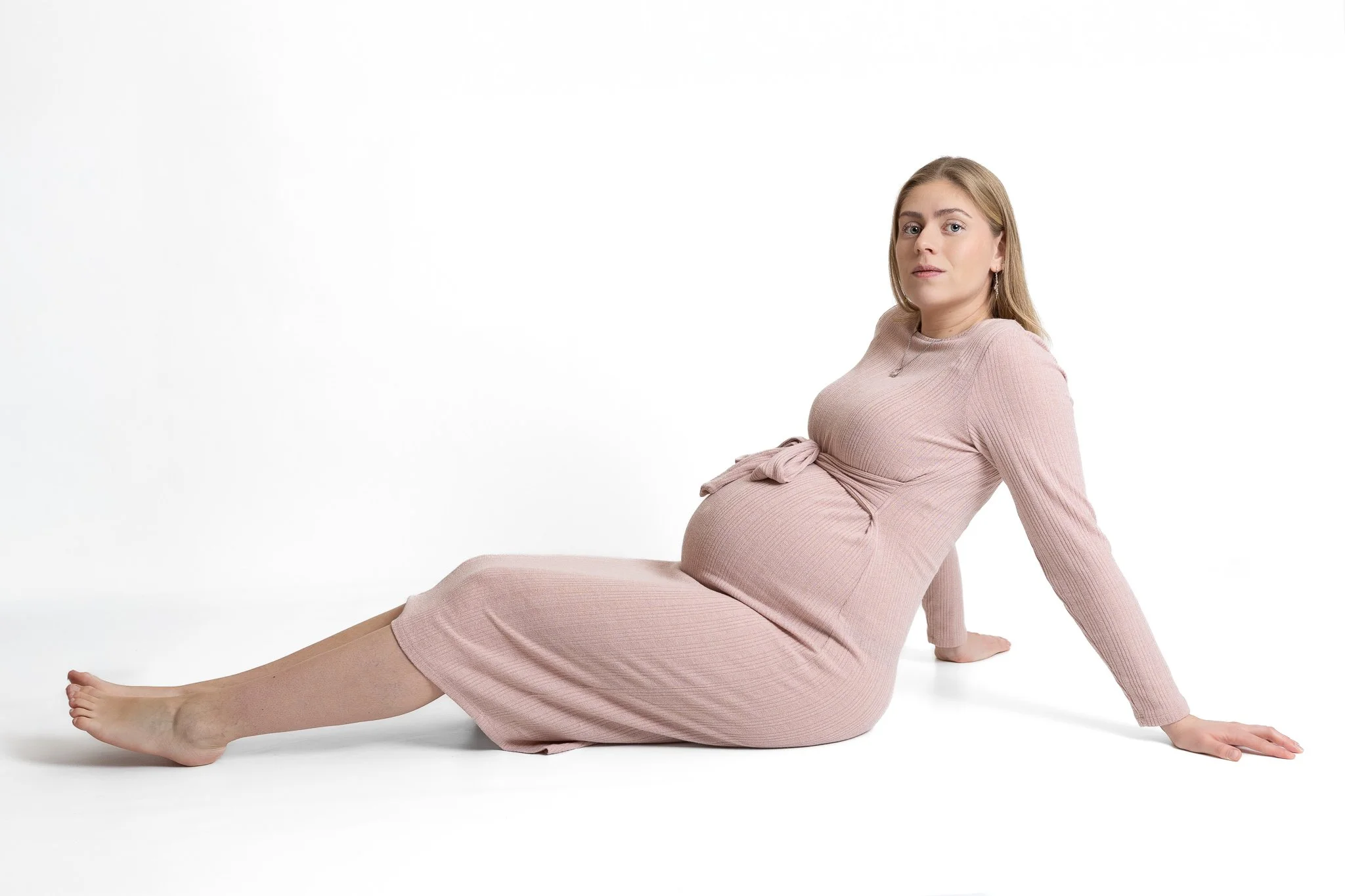 Pregnant woman sitting on the floor with legs extended, wearing a pink dress, against a white background.