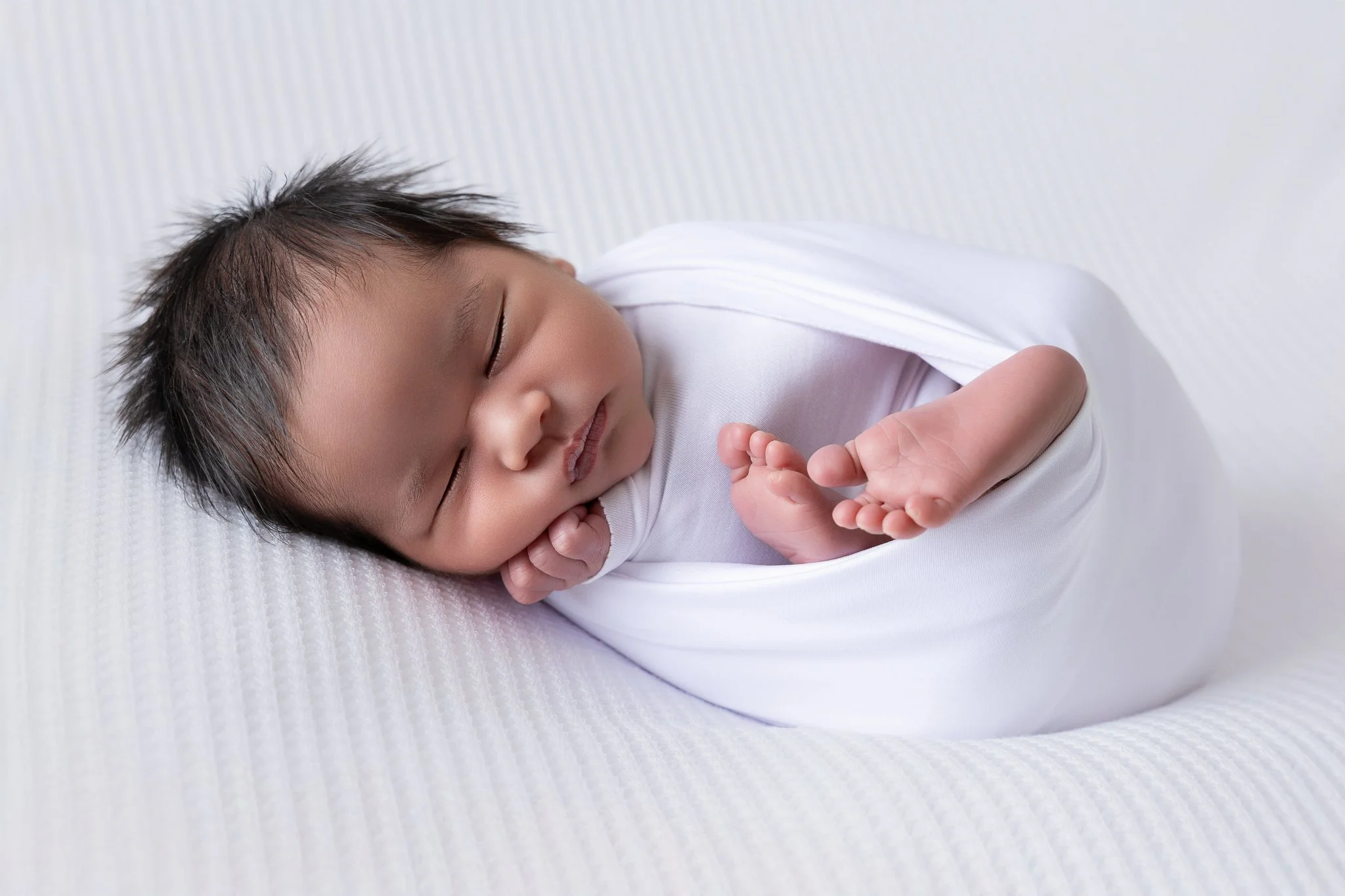 A sleeping baby with dark hair, wearing a white outfit, lying on a soft white textured surface.