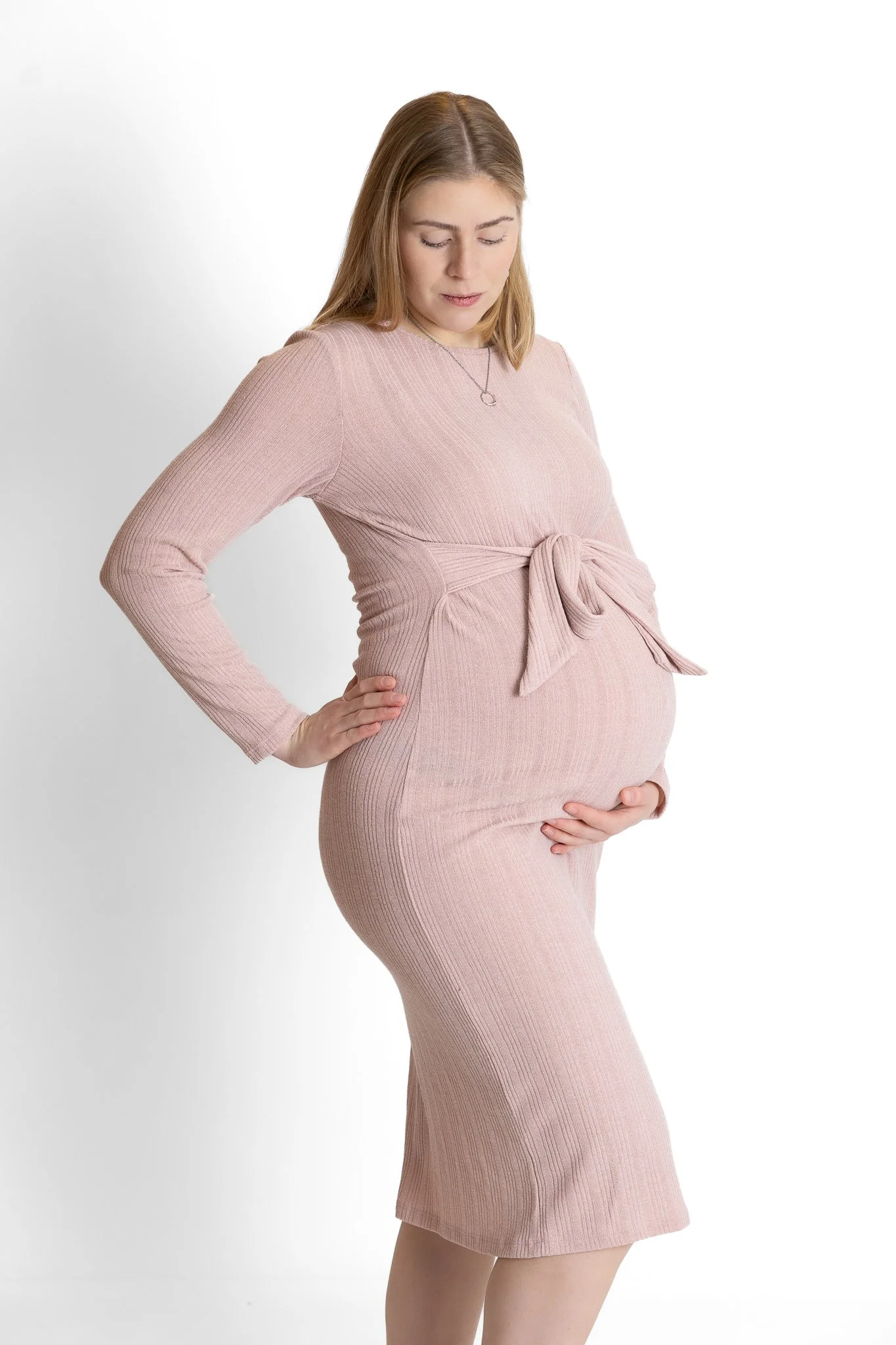 A pregnant woman in a pink, long-sleeved, ribbed dress is standing with one hand on her hip and the other on her belly, looking down and slightly to the side with her eyes closed.