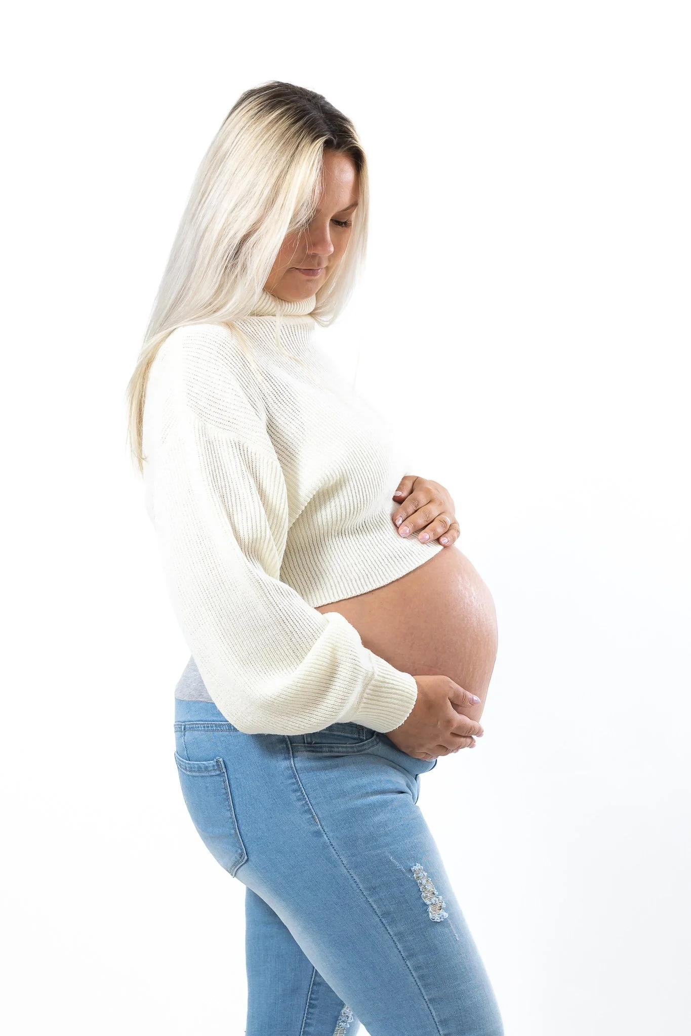 Pregnant woman in a white sweater and jeans holding her belly, standing in profile against a white background.