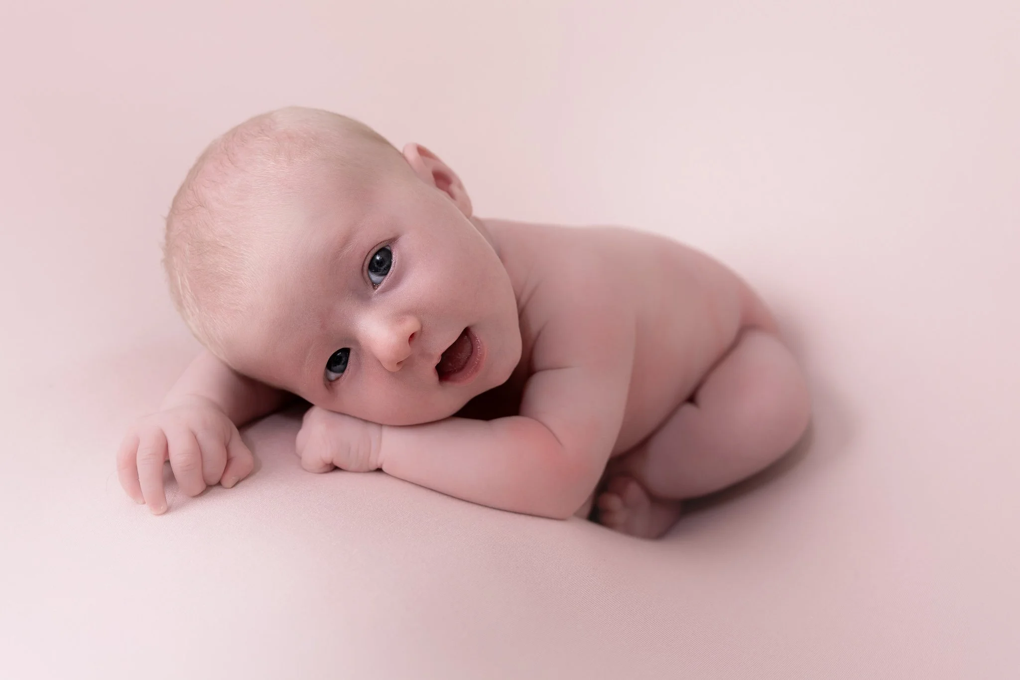 A baby with blue eyes lying on a soft pink surface, looking at the camera with a smiling expression.
