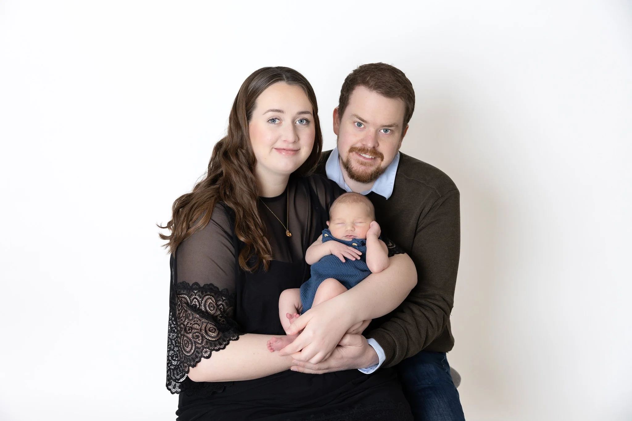A young family of three, with a woman, a man, and a newborn, posing together against a plain white background.
