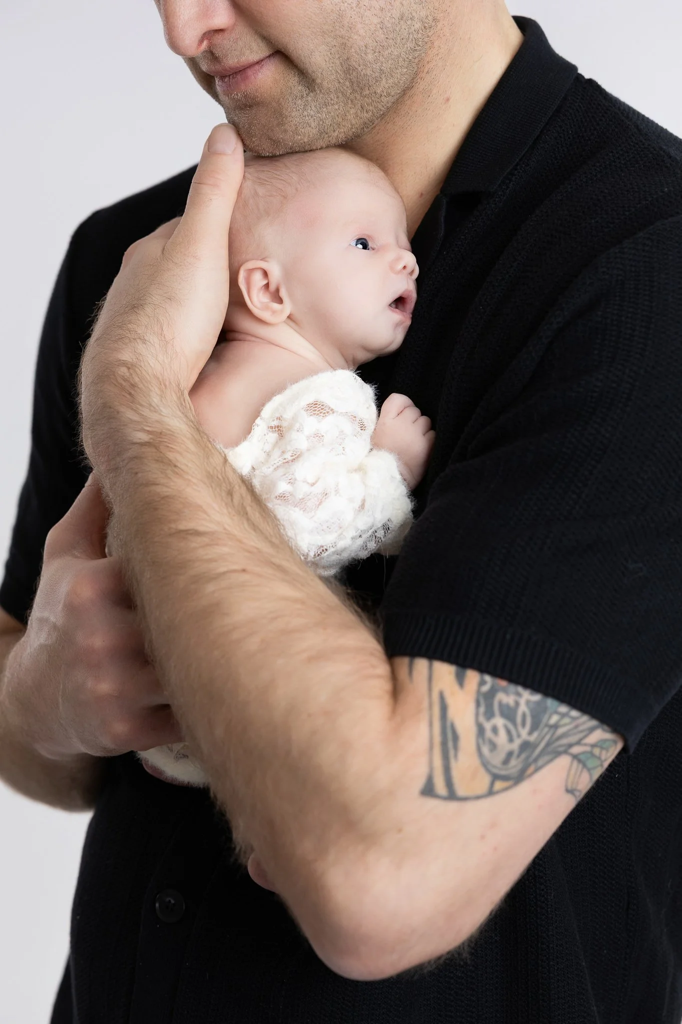 A man with a tattooed arm, short facial hair, and wearing a black shirt is holding a newborn baby close to his chest. The baby has a pacifier in its mouth, light skin, and appears to be sleeping or resting. The man's hand gently holds the baby's head