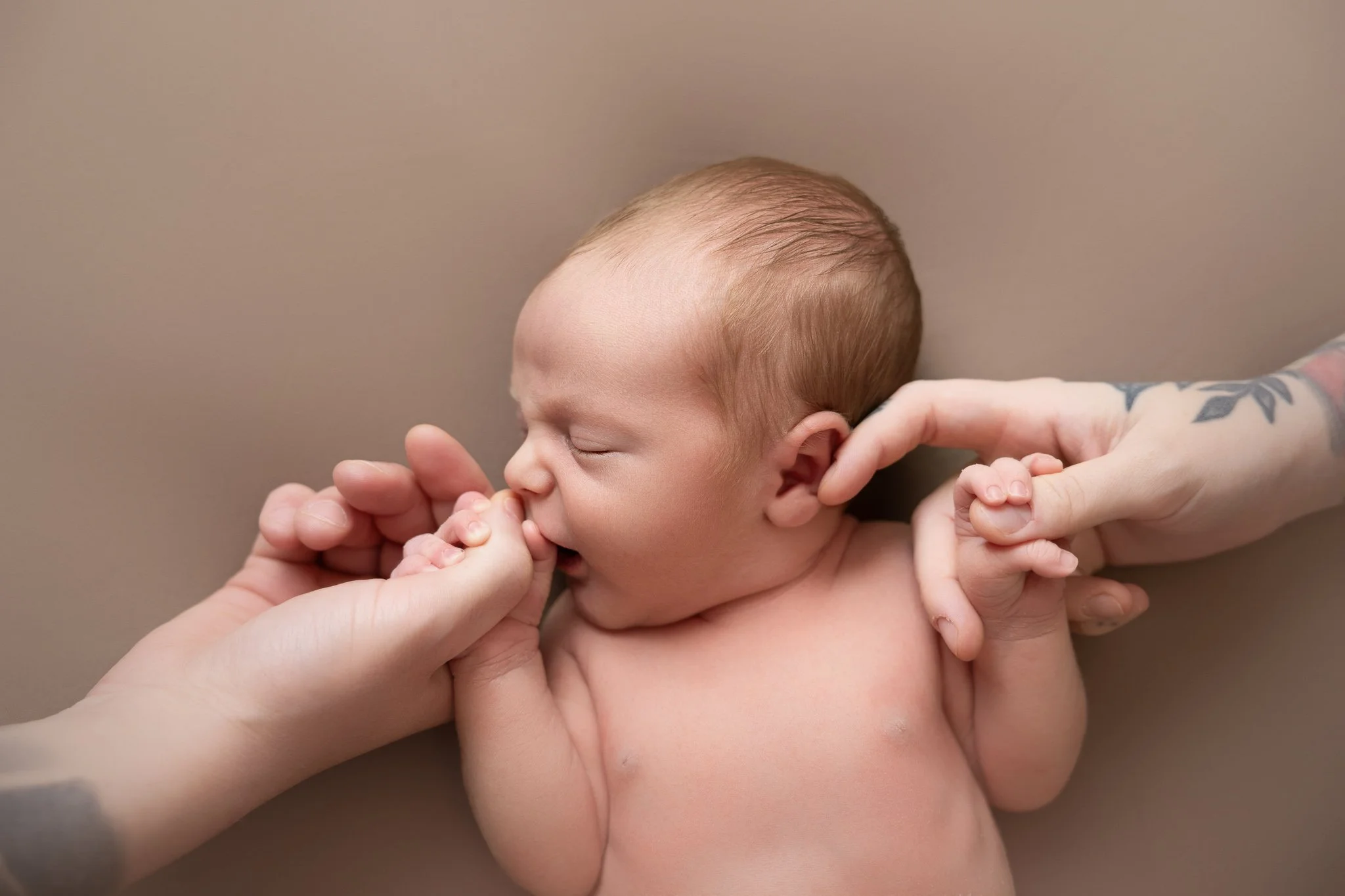 A newborn baby with reddish hair being gently held and touched by adult hands, one hand supporting the baby's head and the other near the face, against a plain beige background.
