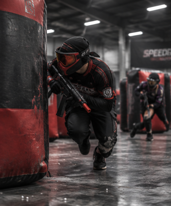Two paintball players crouching behind inflatable barriers in an indoor paintball arena, wearing protective gear including masks and goggles, with one aiming a paintball gun.