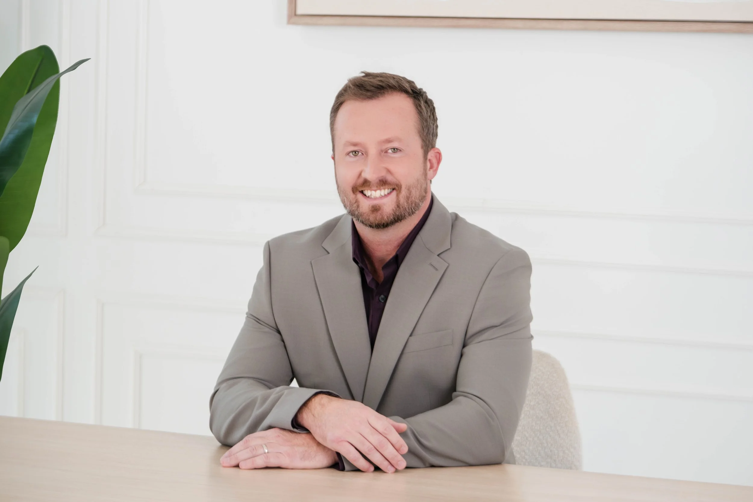 A Caucasian man with a beard and short brown hair, wearing a light gray suit jacket and dark shirt, smiling and sitting at a light-colored table in a bright, white room with a green leafy plant on the left.