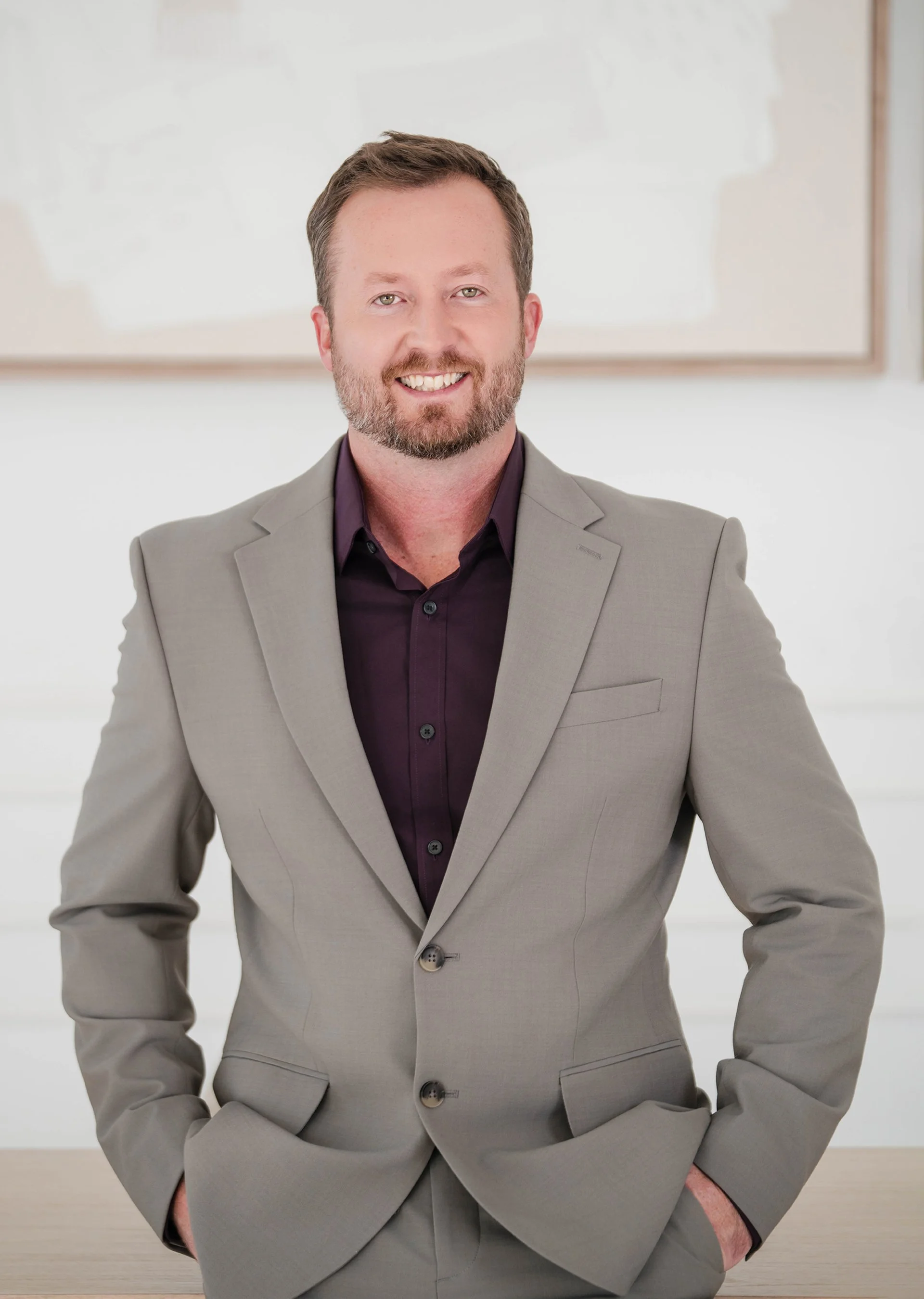 A smiling man with a beard wearing a light gray suit and dark shirt, standing in front of a white background with a whiteboard.