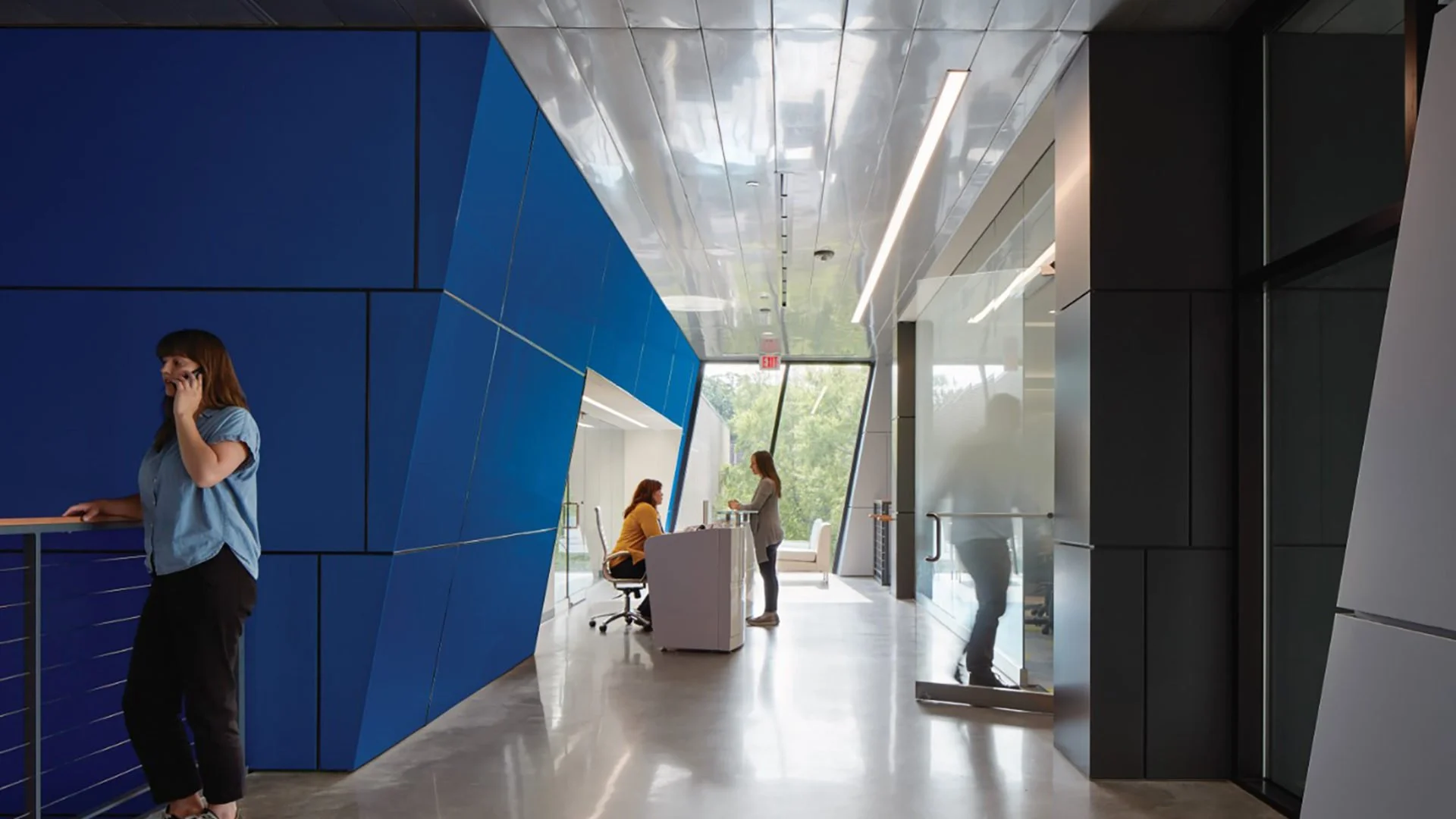 Modern office lobby with blue and black geometric walls, glass doors, and large windows. Two women are seated at a reception desk, and two others are standing and talking, with natural light and greenery visible outside.