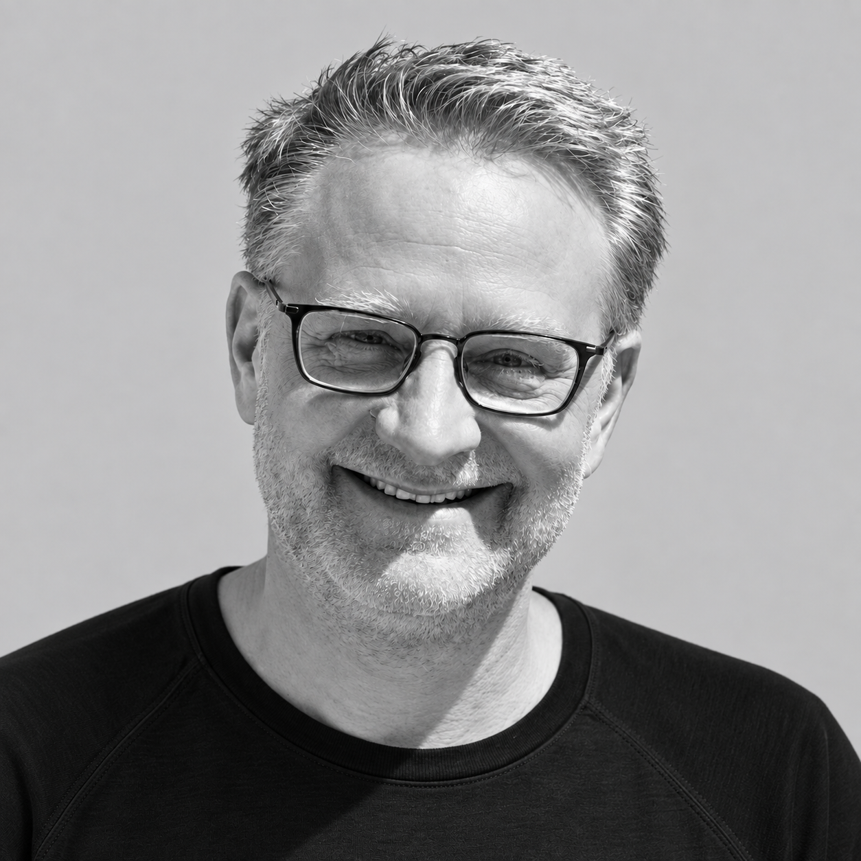 Kenny Beckman - Black and white portrait of a smiling older man with glasses, short gray hair, and a beard, wearing a dark shirt.