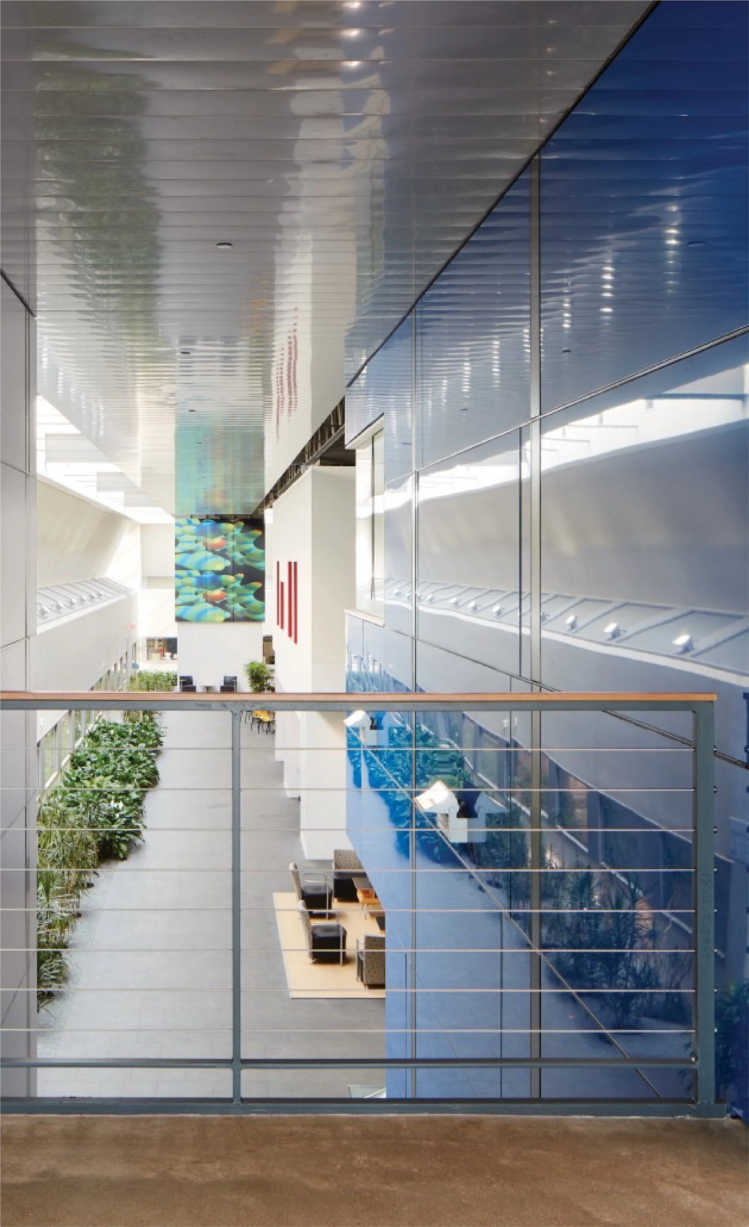 Interior view of a modern building with a balcony overlooking a spacious lobby area. The lobby has seating arrangements, indoor plants, and a large colorful aquatic-themed artwork on the wall. The building features glass walls and a polished floor.