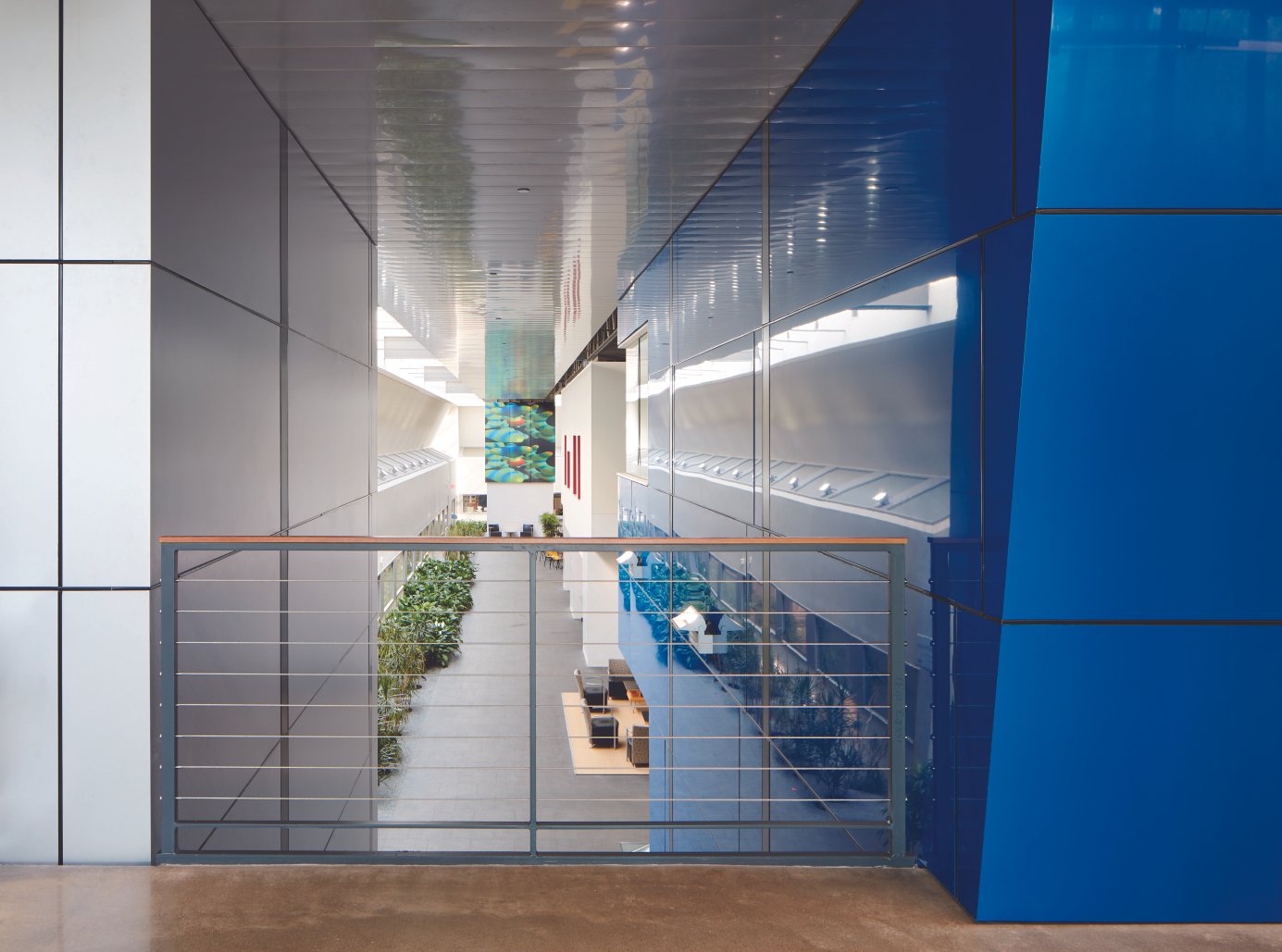 Interior view of a modern, multi-level building with a blue and gray color scheme, featuring a hallway, plants, artworks, and seating areas.