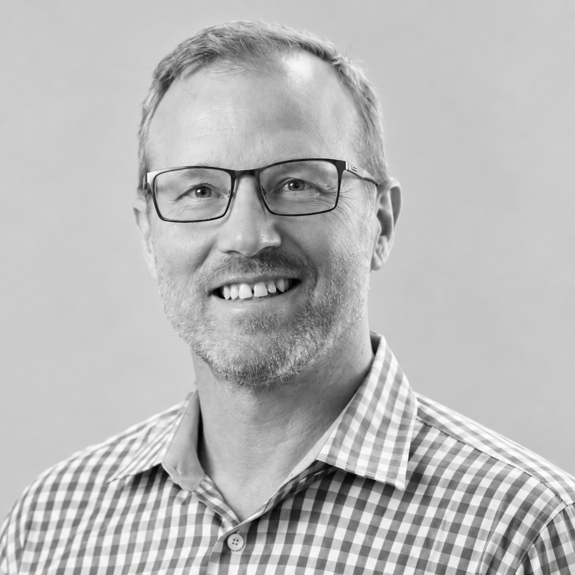 Sam Shuster - A black and white portrait of a smiling man wearing glasses and a checkered shirt.