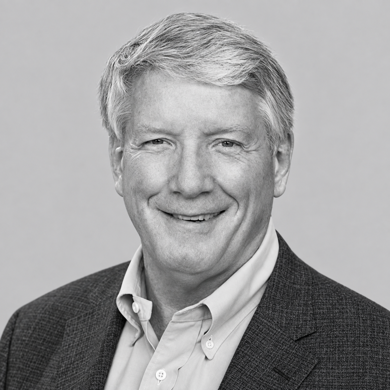 Black and white headshot of a smiling middle-aged man with light-colored hair, wearing a collared shirt and blazer against a plain background.