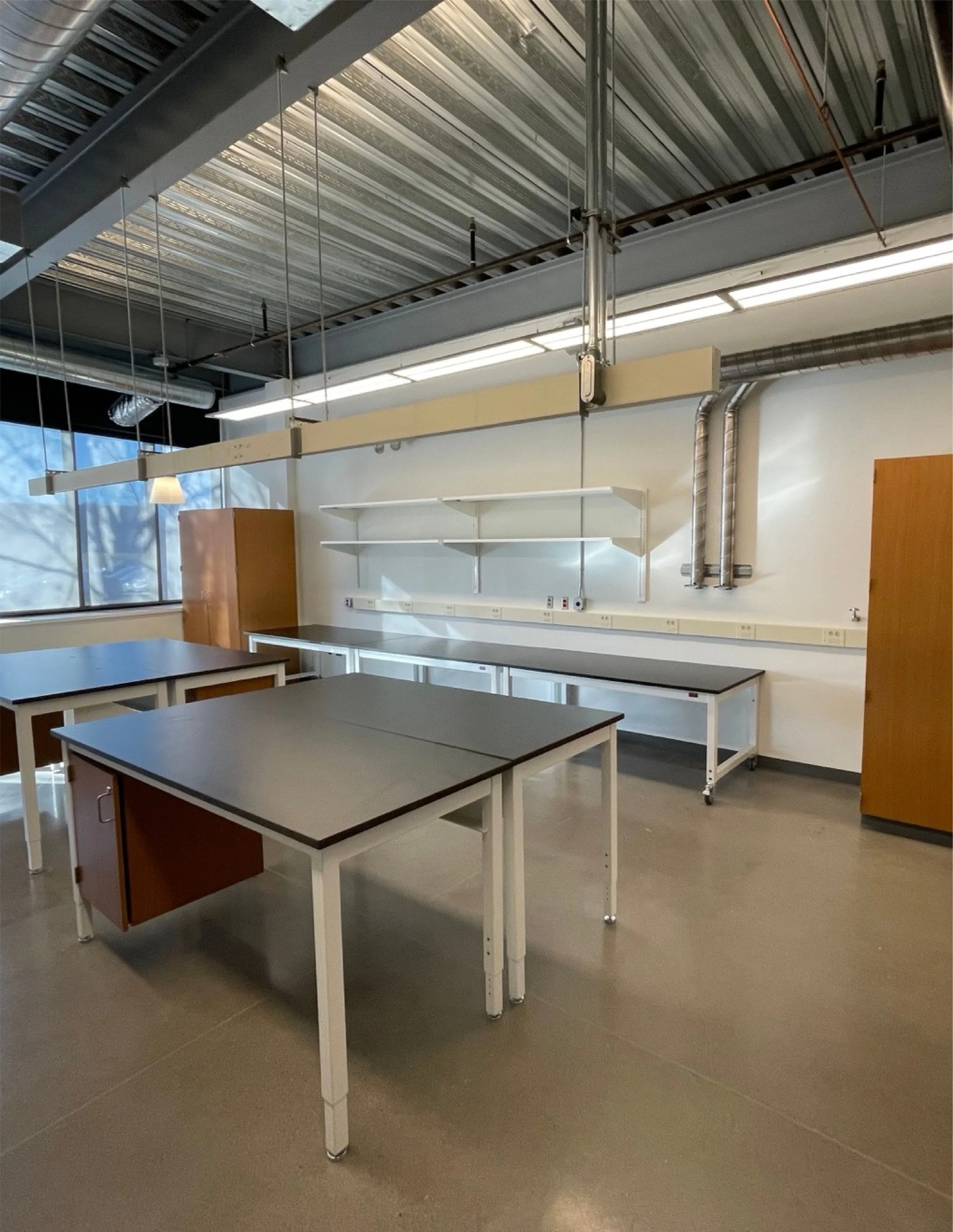 Empty classroom or laboratory with tables, shelves, electrical outlets, exposed ductwork, and industrial lighting.