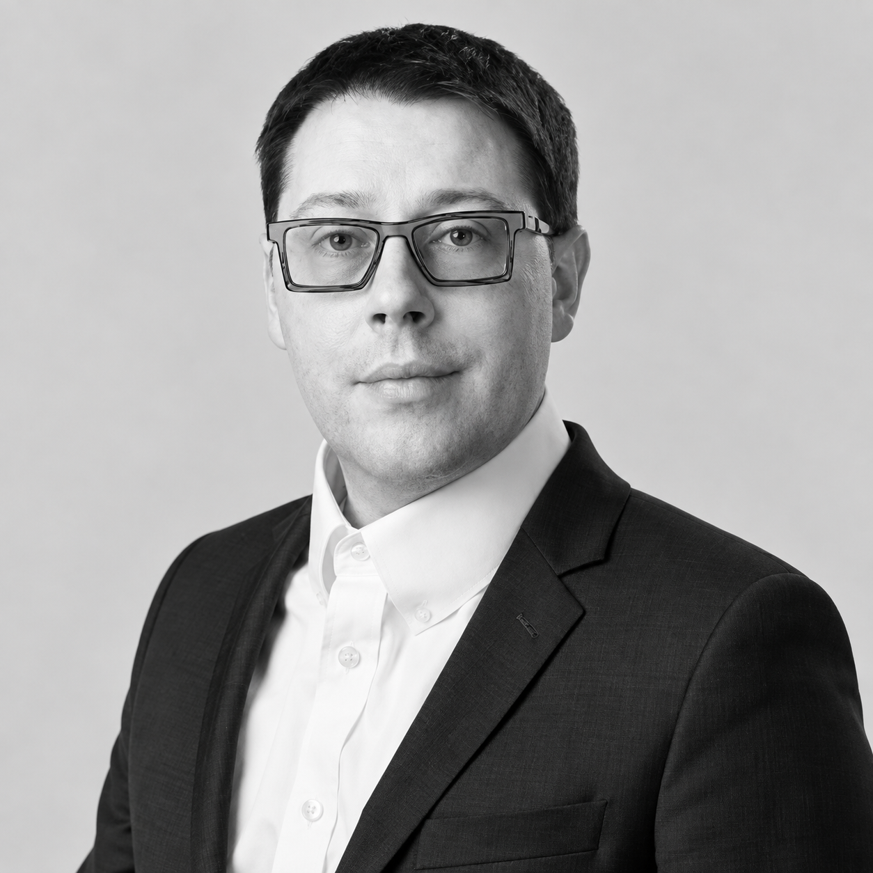 Frank Jaskulke - Black and white professional headshot of a man wearing glasses, a white collared shirt, and a dark suit blazer.