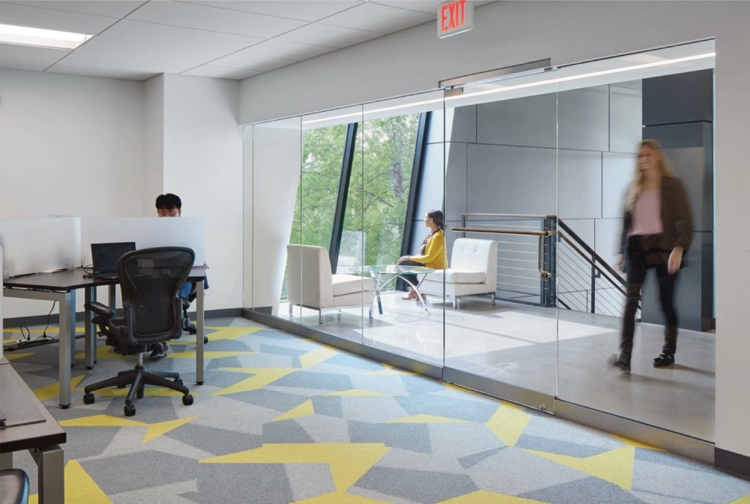 Modern office space with glass partition, people working and walking, colorful carpet, large window with outdoor trees, and an illuminated exit sign.