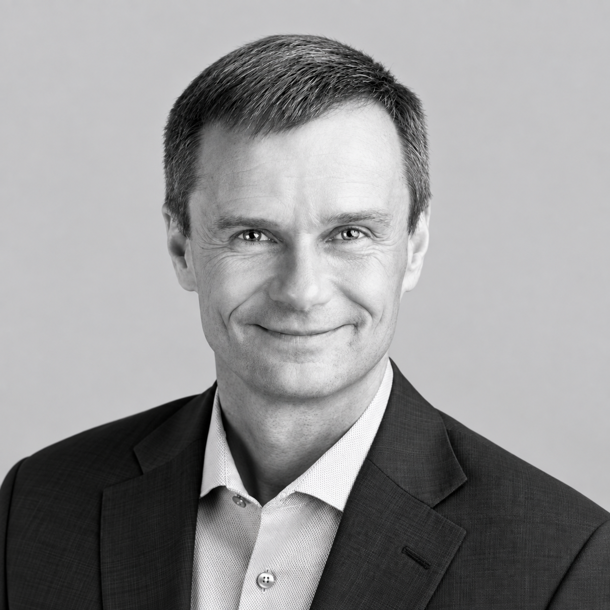 Paul Owen - Black and white professional portrait of a smiling man in a suit and collared shirt.
