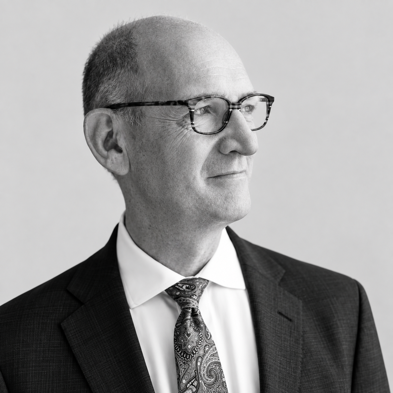 Cris Ross - Black and white portrait of a middle-aged man with glasses, wearing a suit and patterned tie, looking to his right with a thoughtful expression.