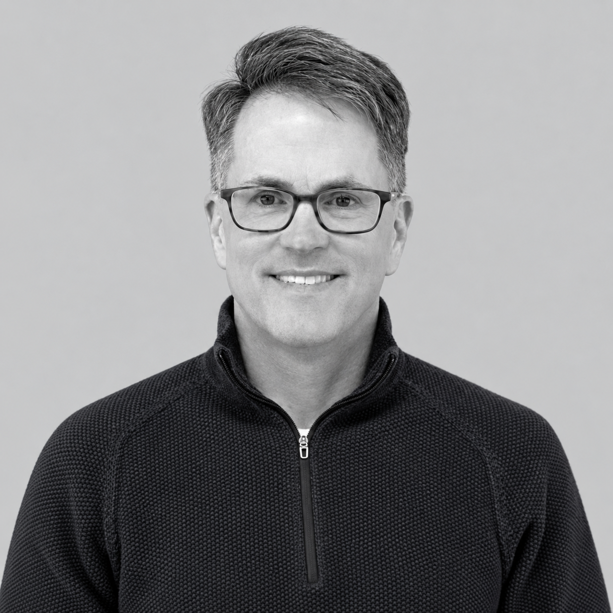 Jack Starr - Black and white portrait of a smiling man wearing glasses and a zip-up textured sweater, standing against a plain background.