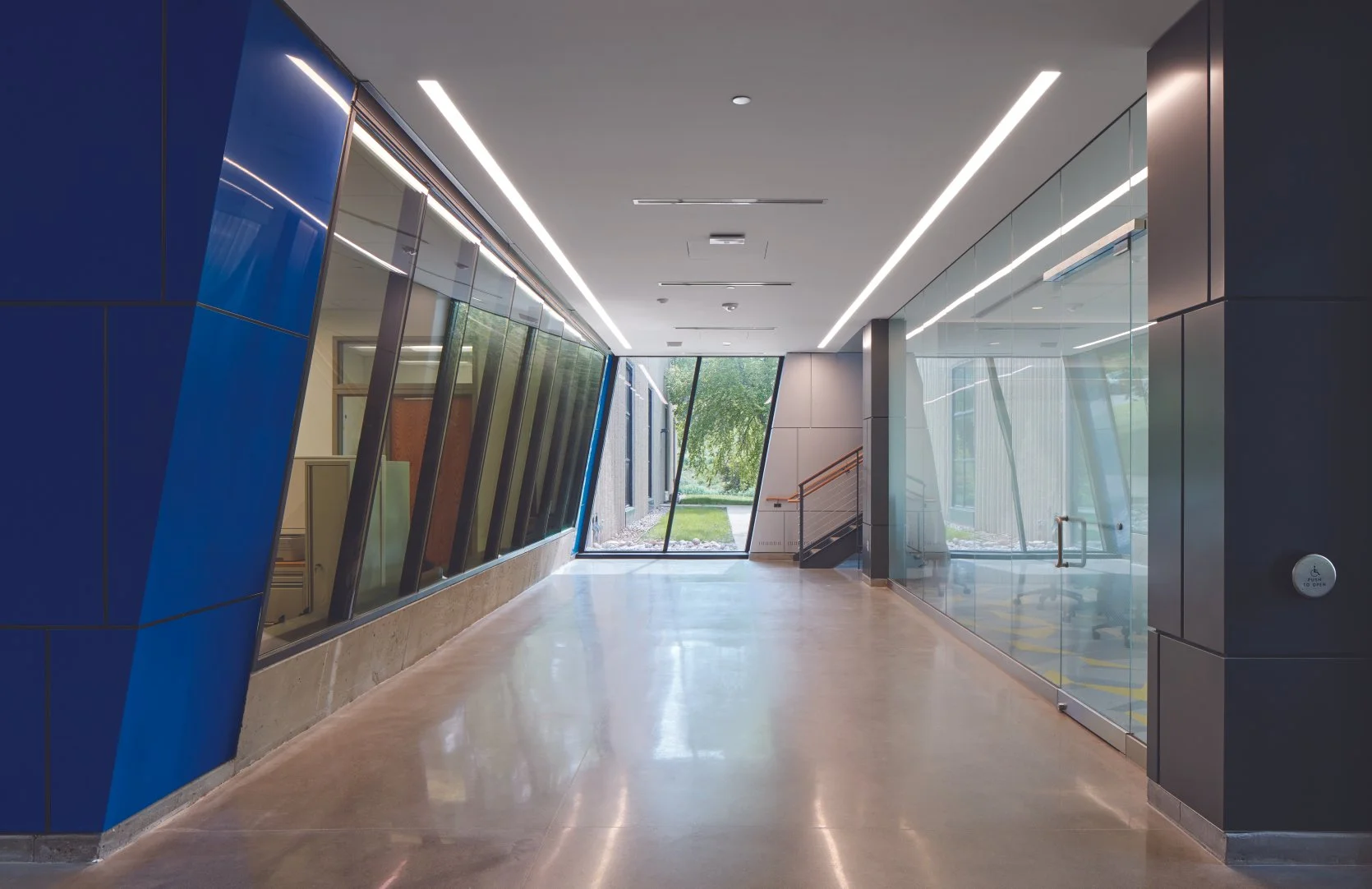Modern hallway with large glass windows on both sides, wood and metal accents, and a view of green trees outside.