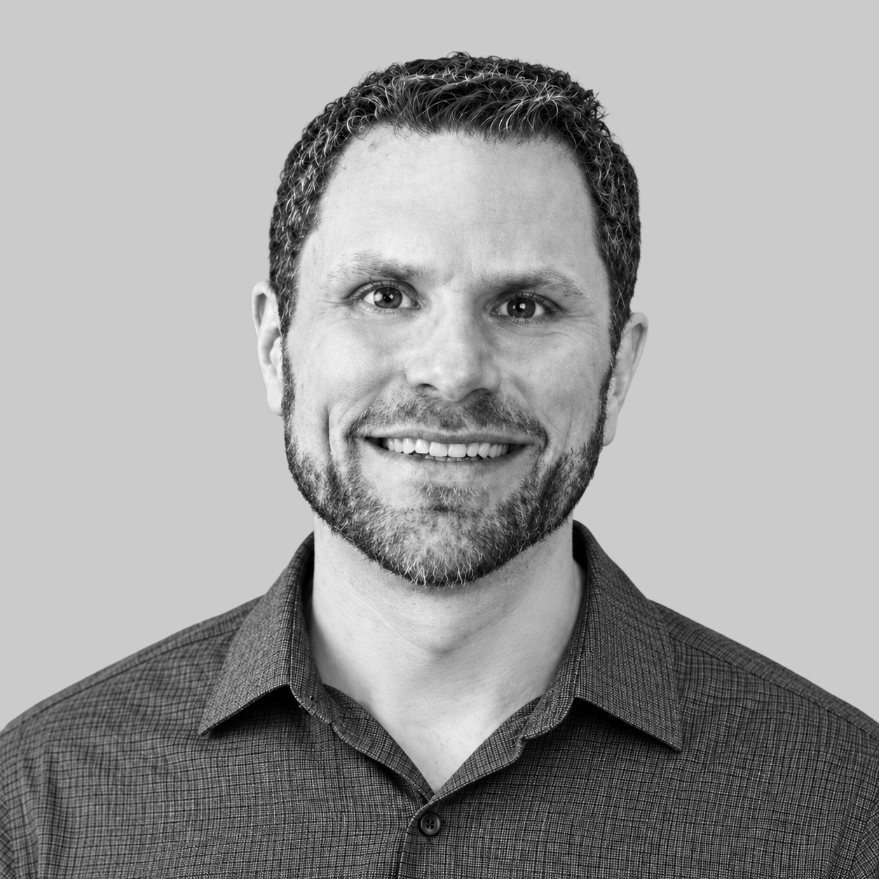 Chase Orsello - A smiling man with short wavy hair, beard, and mustache, wearing a patterned button-up shirt, against a plain background.