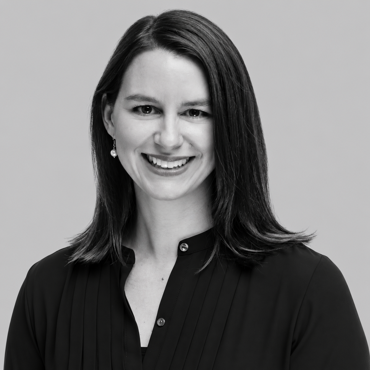 Jessica Mogilka - Black and white portrait of a smiling woman with shoulder-length dark hair, wearing a button-up shirt and earrings.